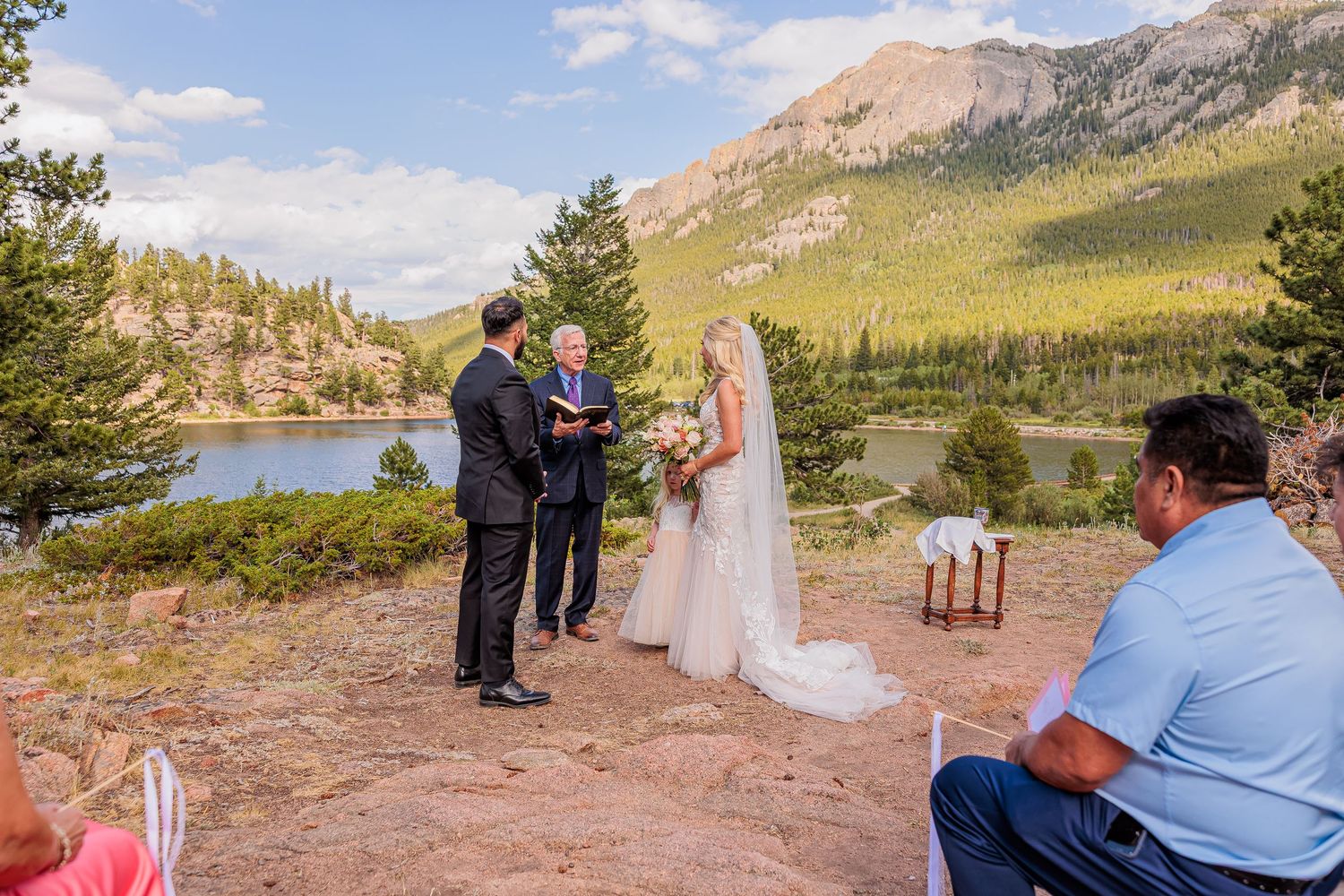 Outdoor wedding ceremony with couple exchanging vows on rocky overlook with mountain lake and forest backdrop.