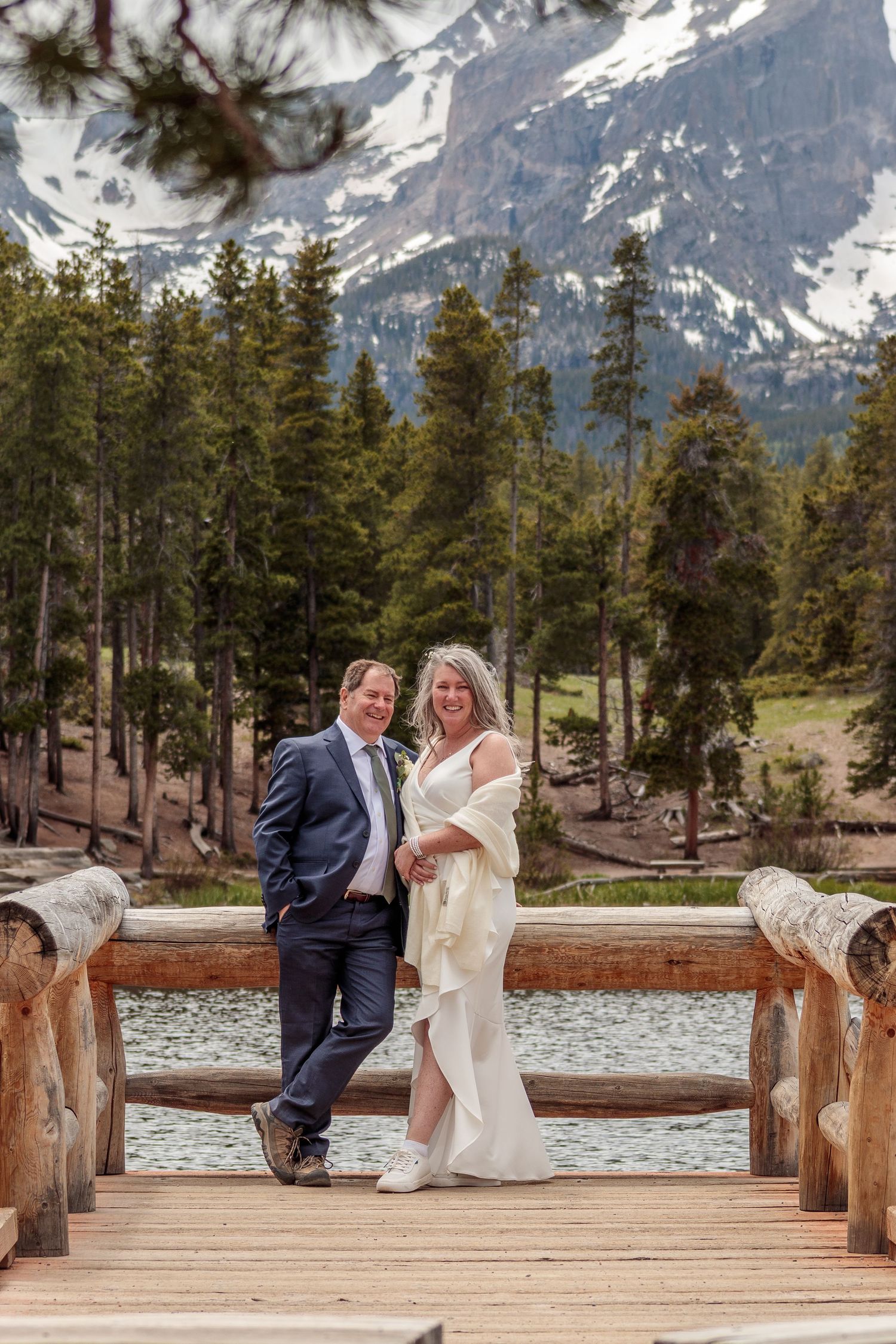 Couple sitting together on wooden dock with snowy mountain peaks and evergreen forest in background.