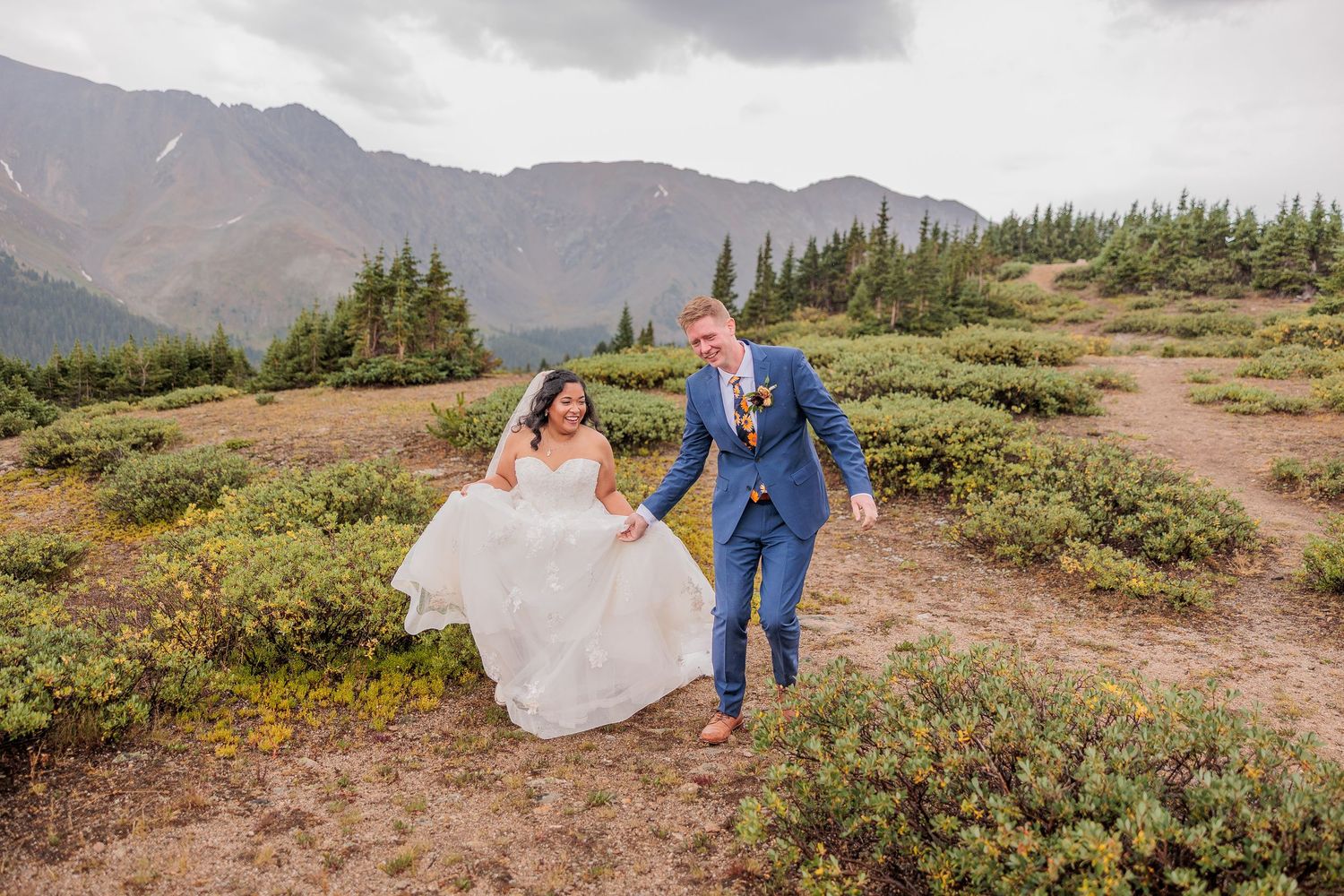 A couple walks along a scenic mountain trail with dramatic cloudy skies and wildflowers in the Colorado landscape.