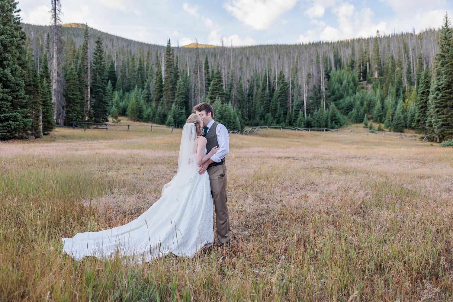 A couple in wedding attire embraces in a mountain meadow surrounded by evergreen trees and golden grass.