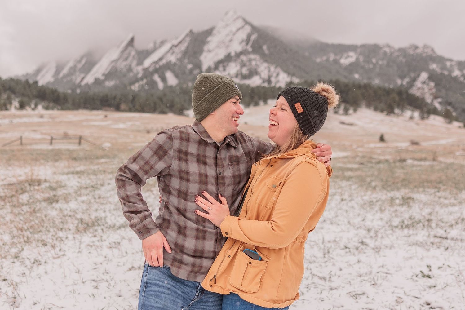 A couple embraces in a snowy mountain landscape wearing plaid and orange winter clothing.
