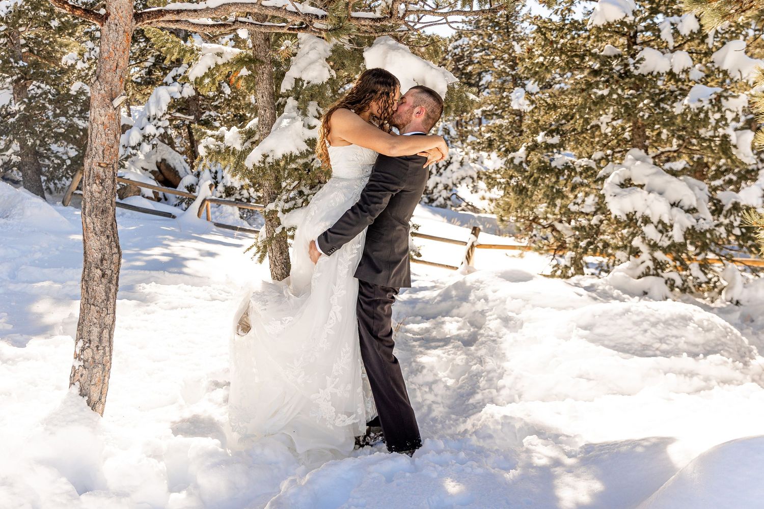 A couple in wedding attire embrace in a snowy winter forest setting with sunlight filtering through the trees.