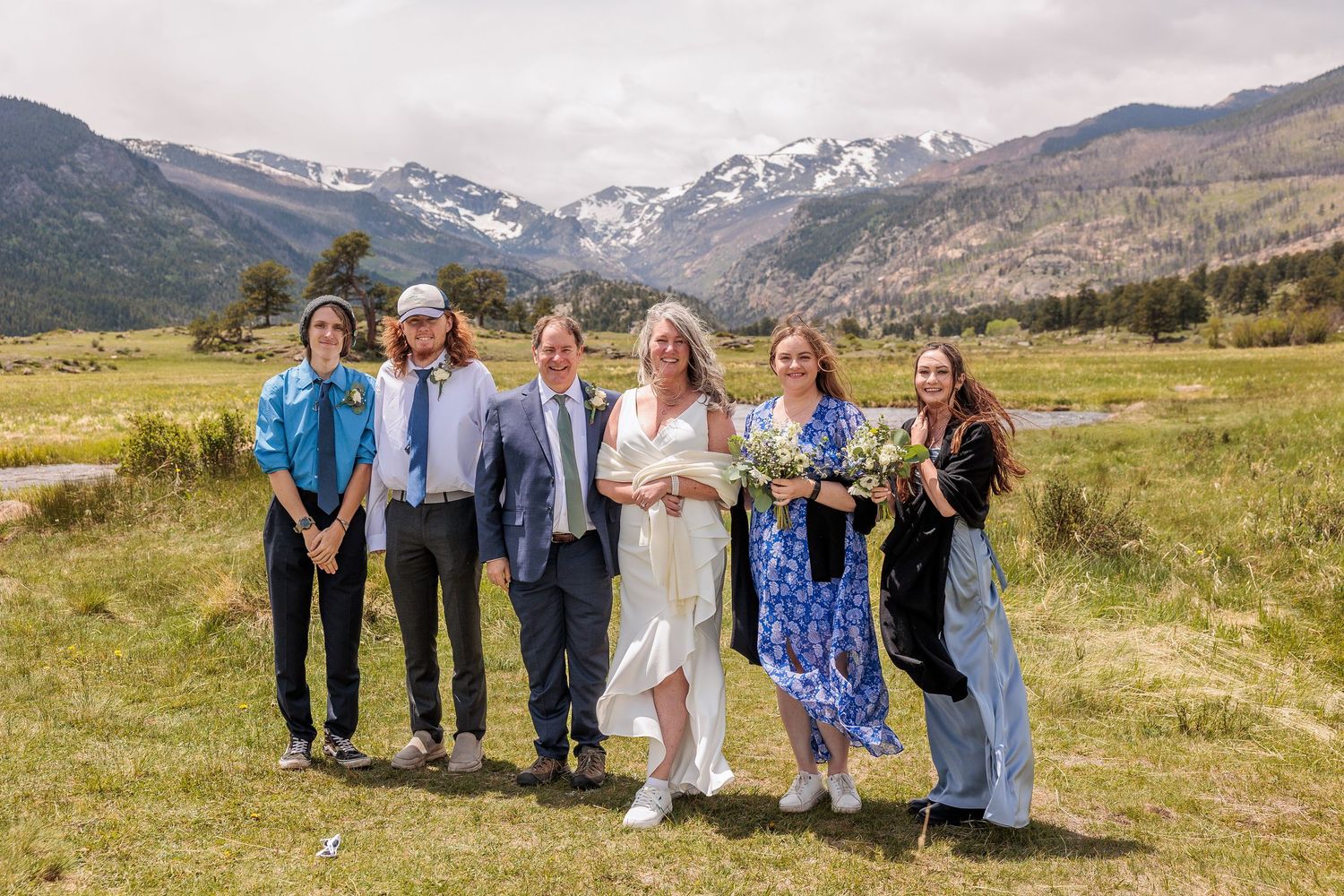 A wedding party poses together in a mountain meadow with snow-capped peaks in the background.