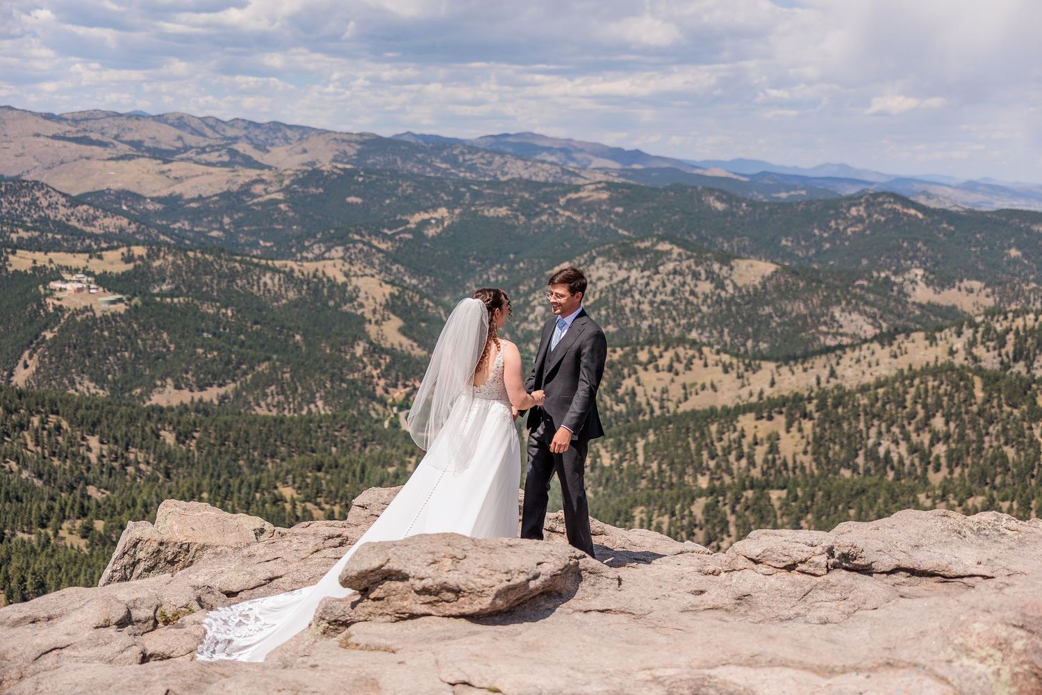 A couple stands on a rocky mountain overlook with sweeping views of forested peaks and valleys in the distance.