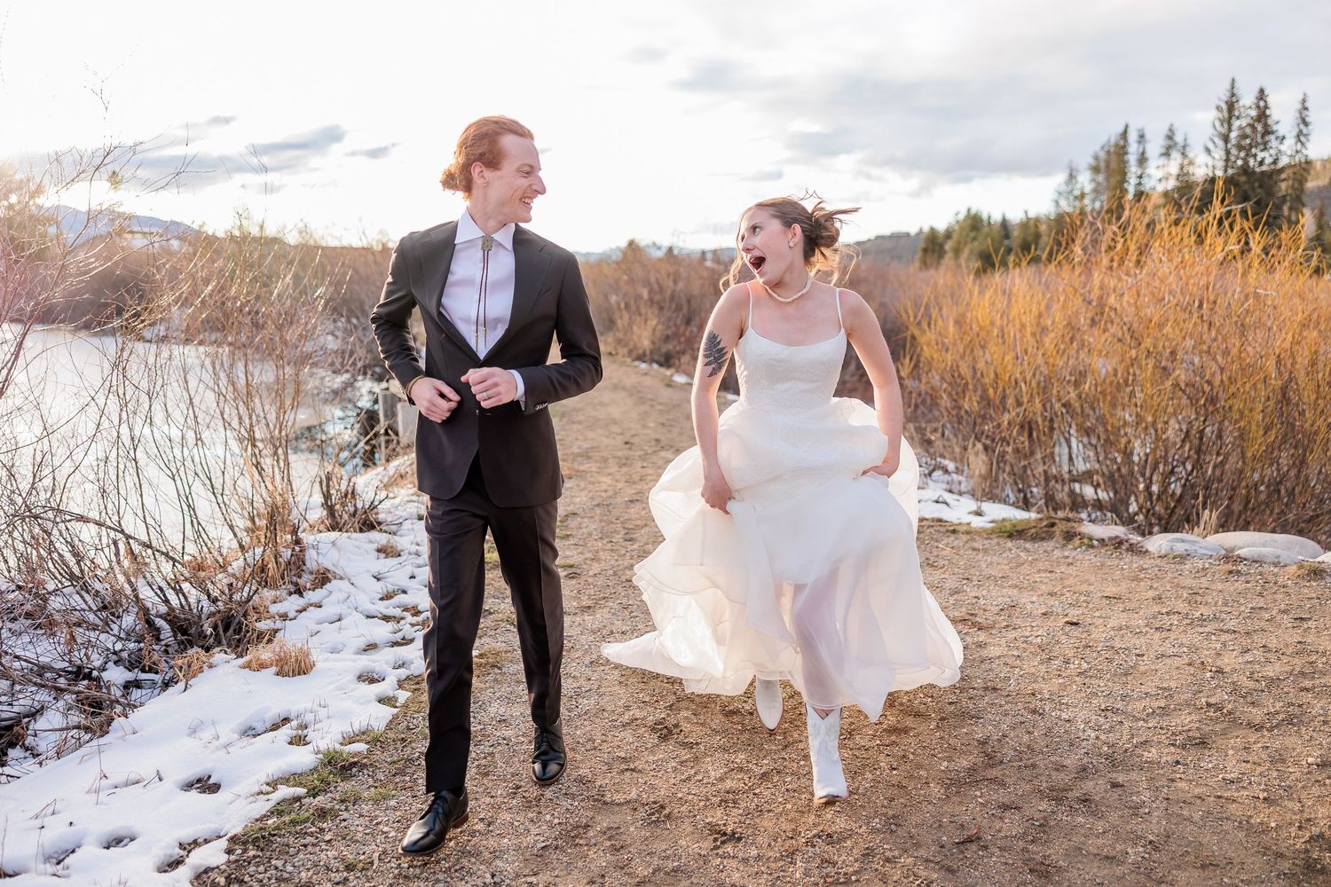 A couple in wedding attire walks along a snowy path through a winter landscape at sunset.