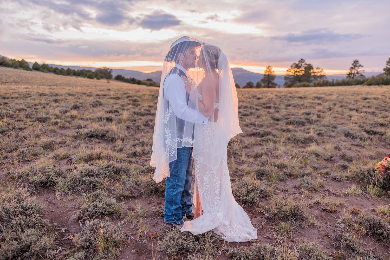 Couple in white and denim embrace during golden hour in a rustic desert landscape with mountains in the background in a National Park