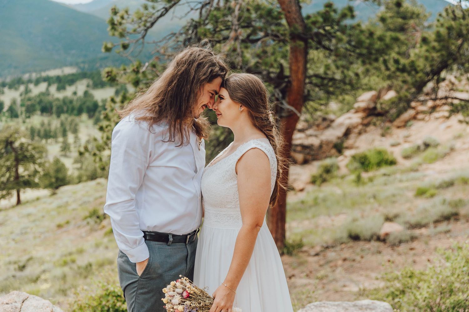 A romantic mountain wedding photo with a couple embracing on a scenic hillside overlooking a forested valley.