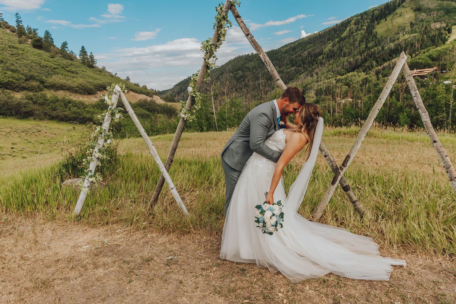 A romantic mountain wedding ceremony with a wooden arch and white fabric decor set against scenic rolling hills.