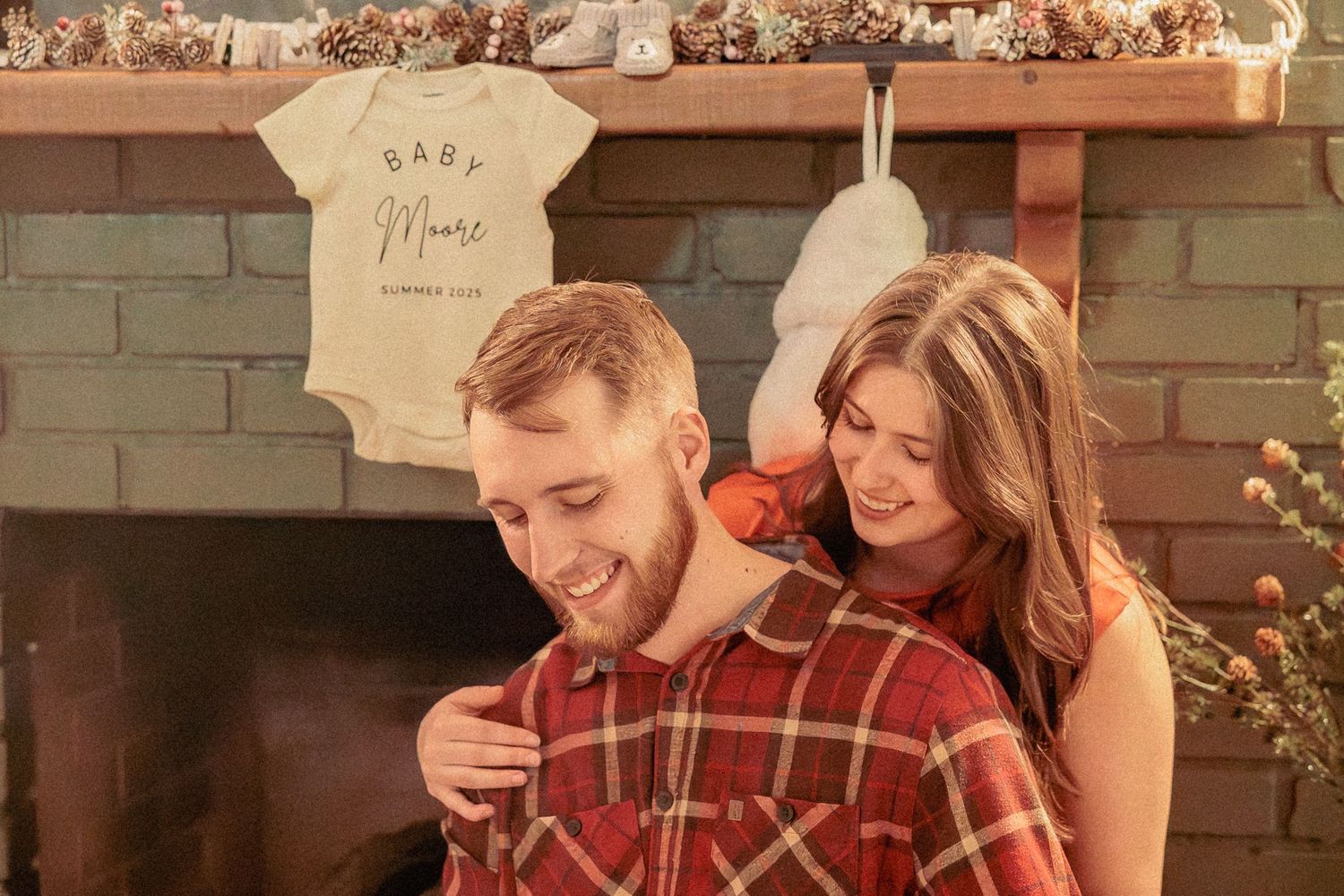 A couple shares a joyful moment together in front of a brick fireplace with a baby onesie hanging in the background.