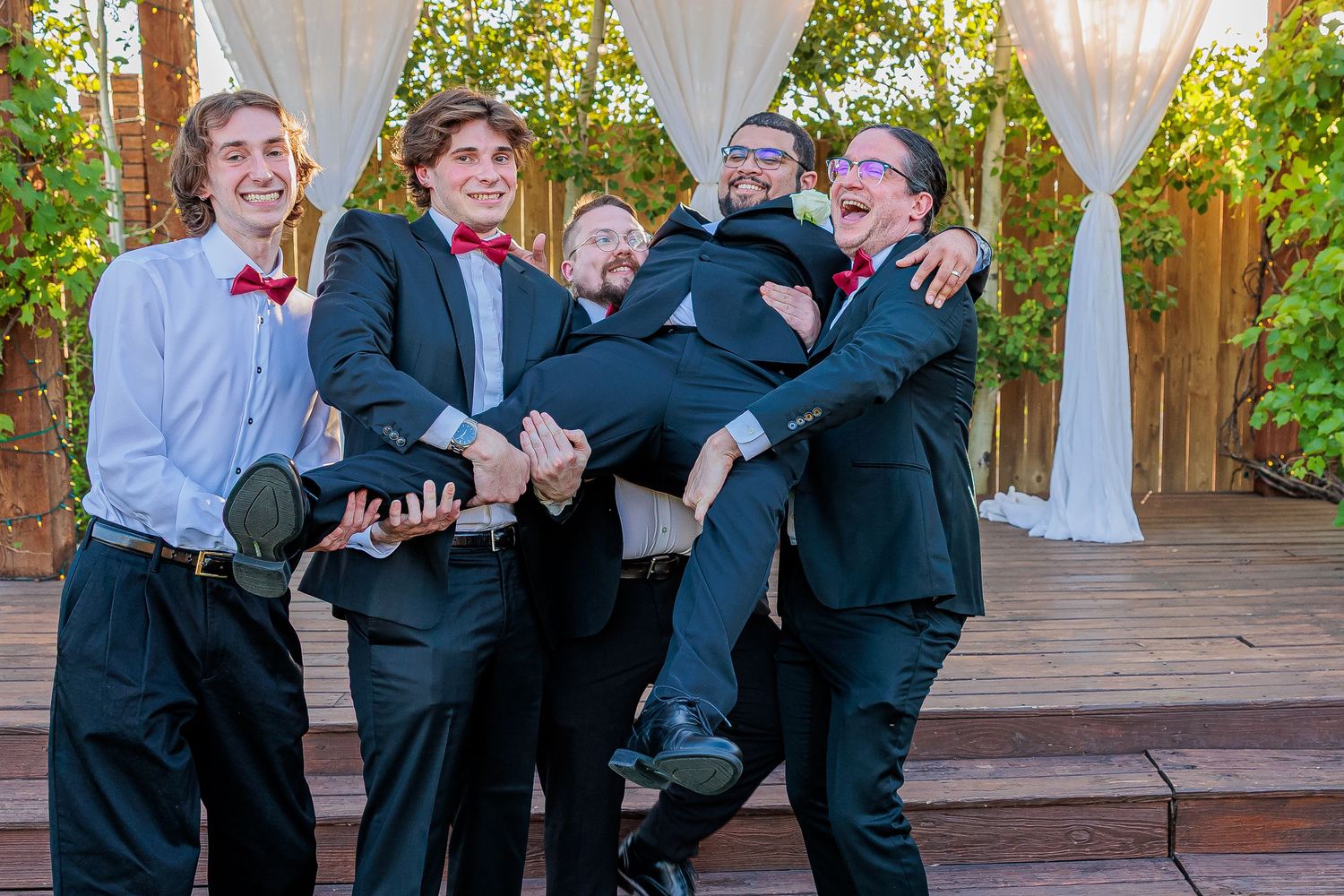 Groomsmen in black suits and red bow ties playfully lifting the groom outdoors at a wedding venue.