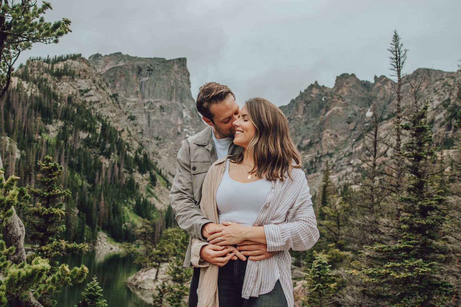 A couple shares an intimate moment in the mountains surrounded by evergreen trees and dramatic rocky peaks.