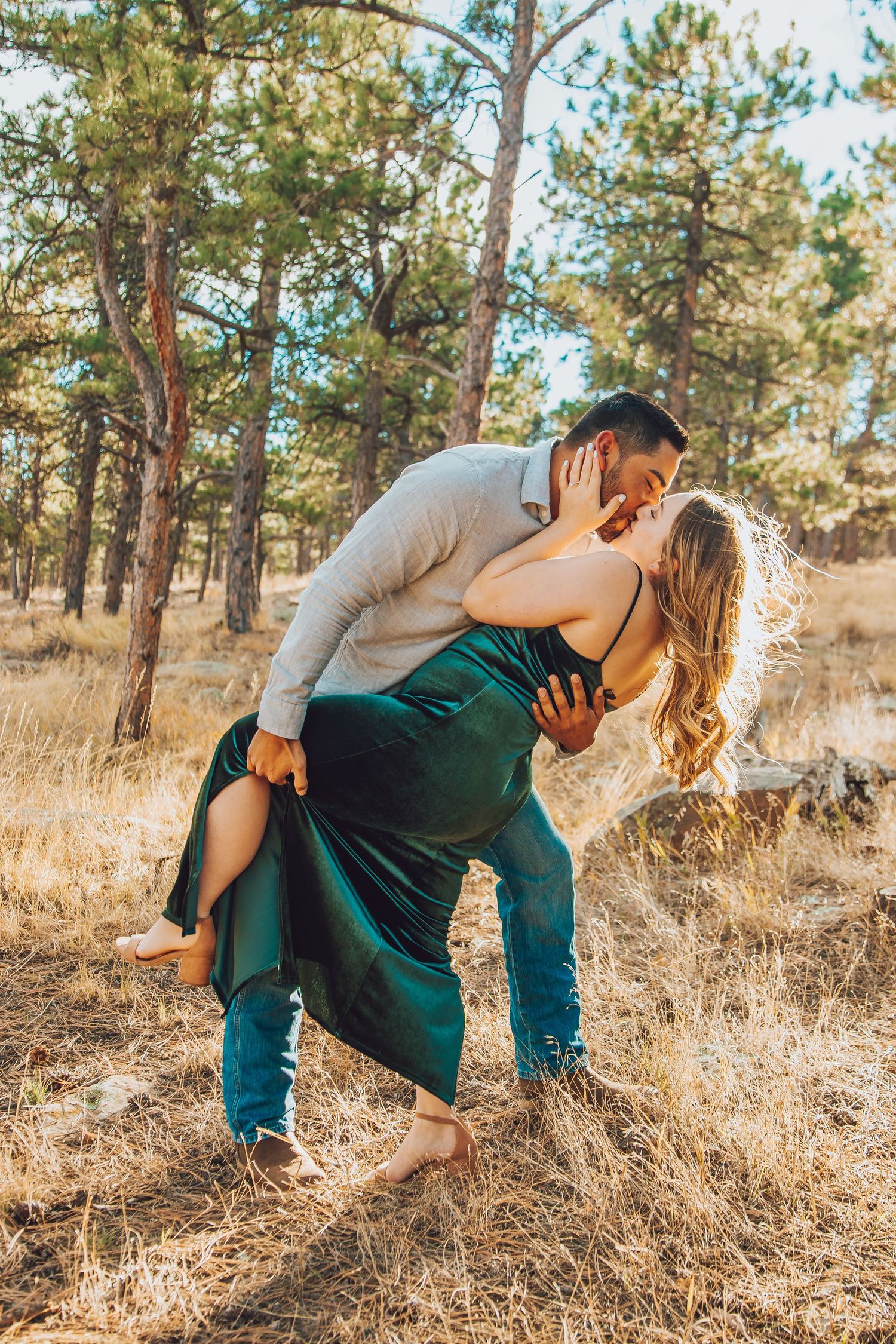 Couple sharing a romantic dip kiss in a sunlit pine forest, man in gray shirt, woman in emerald green dress.
