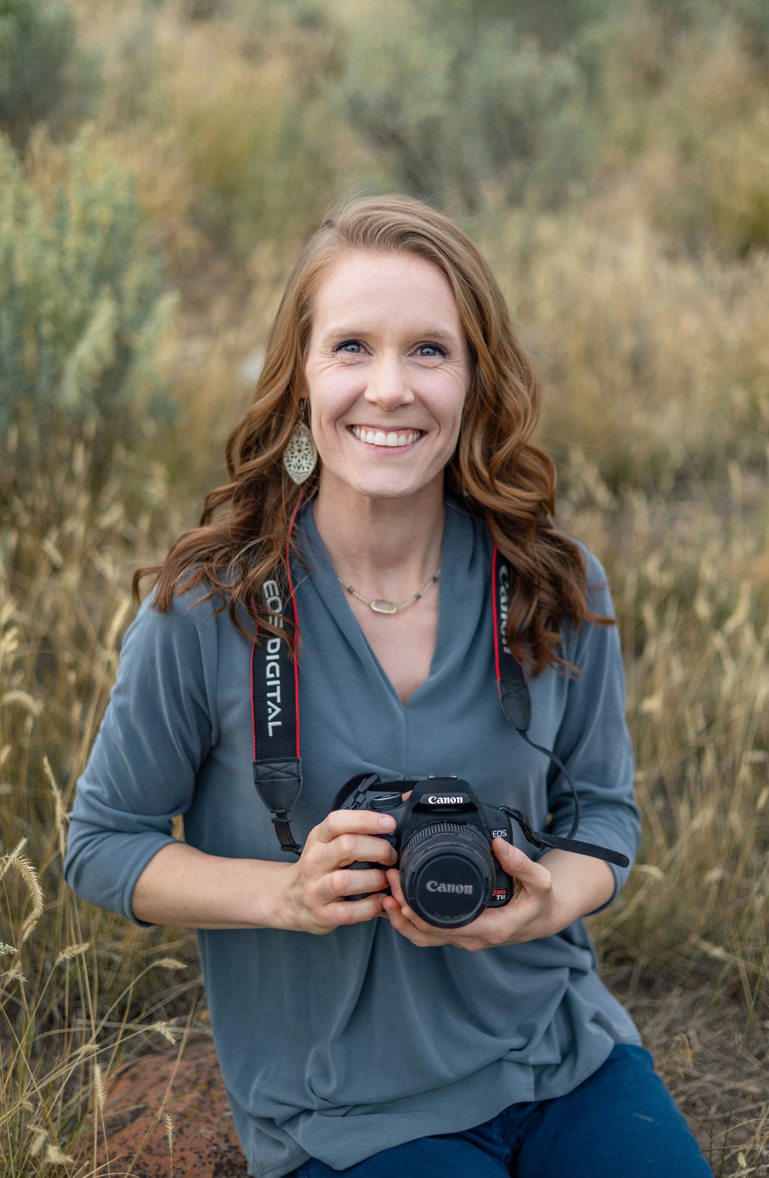 Smiling woman photographer holding a Canon DSLR camera outdoors in a field, wearing a gray top and Canon camera strap.