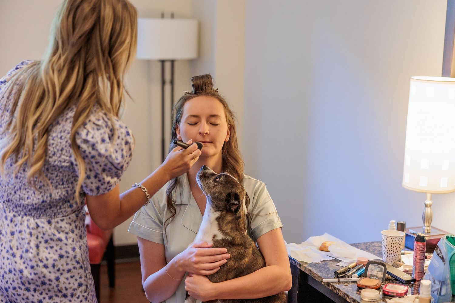Someone gets makeup applied while holding a small dog in a dressing room with lamps.