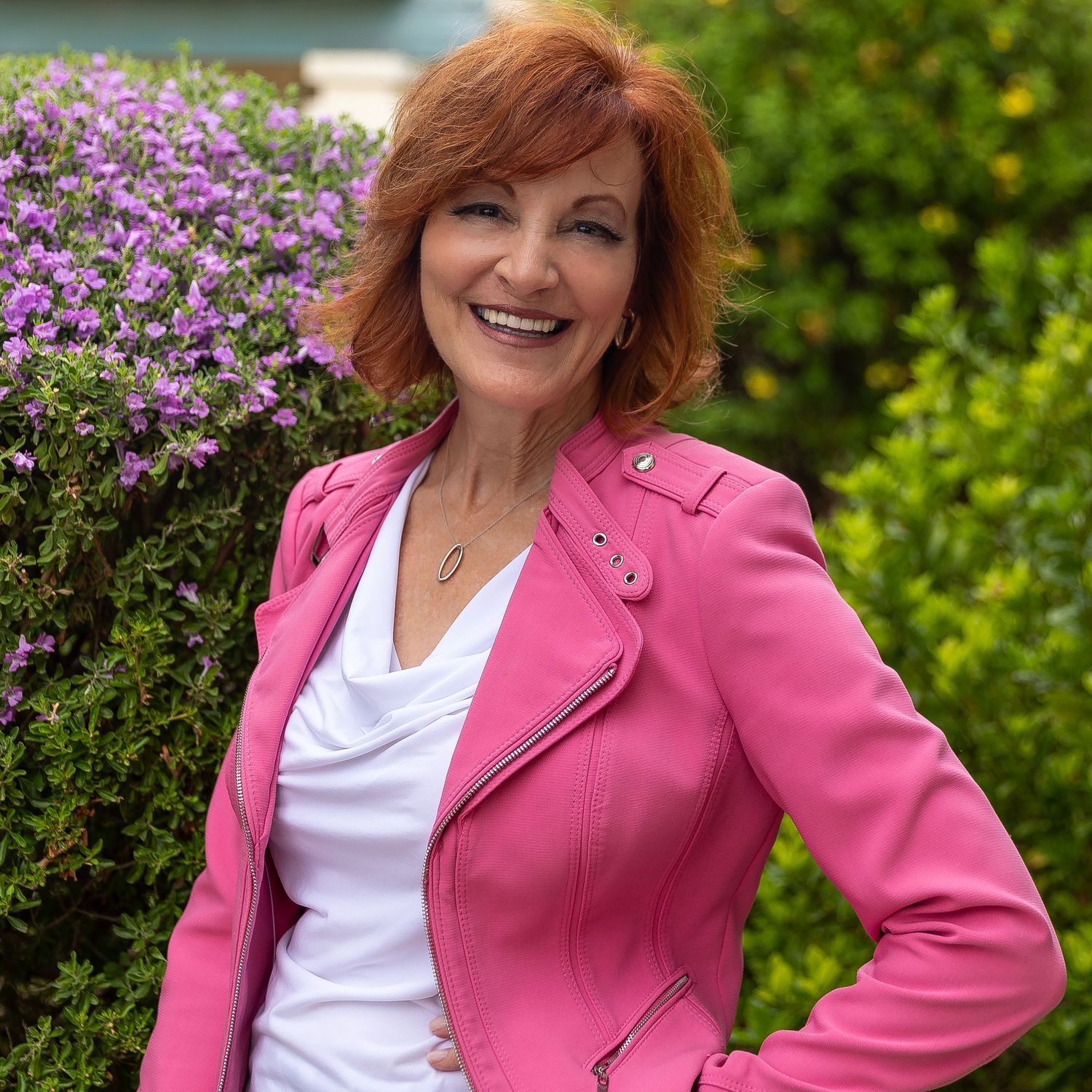 Someone wearing a bright pink leather jacket and white top poses against a backdrop of purple flowers and greenery.