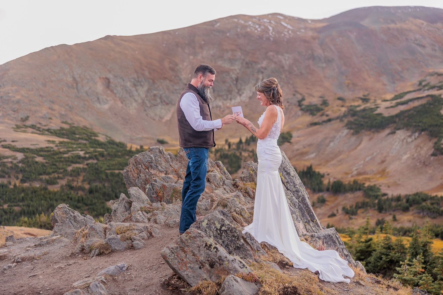 Wedding couple standing on rocky mountain ledge with scenic mountain backdrop.