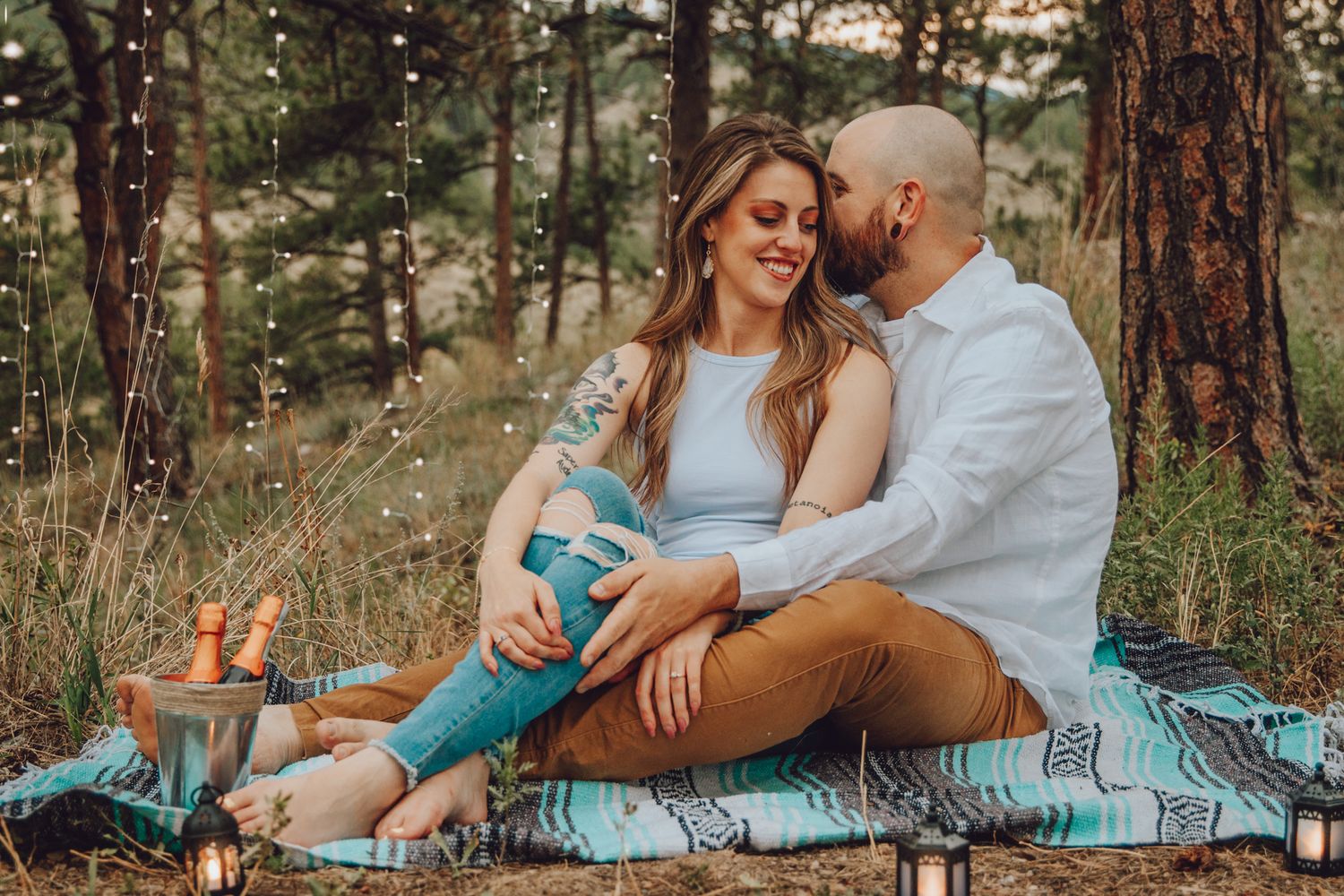 A couple sits together on a turquoise blanket in a sunlit pine forest during a romantic outdoor photoshoot.