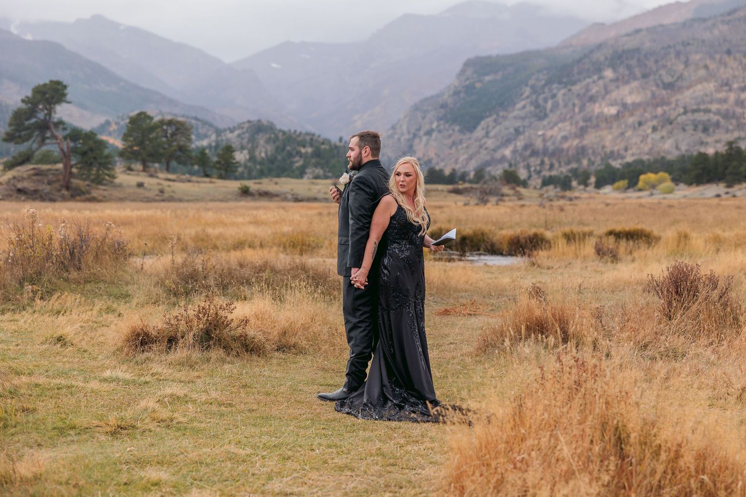 A couple in formal attire stands in a scenic mountain meadow with golden grass and rugged peaks in the background at Morraine Valley