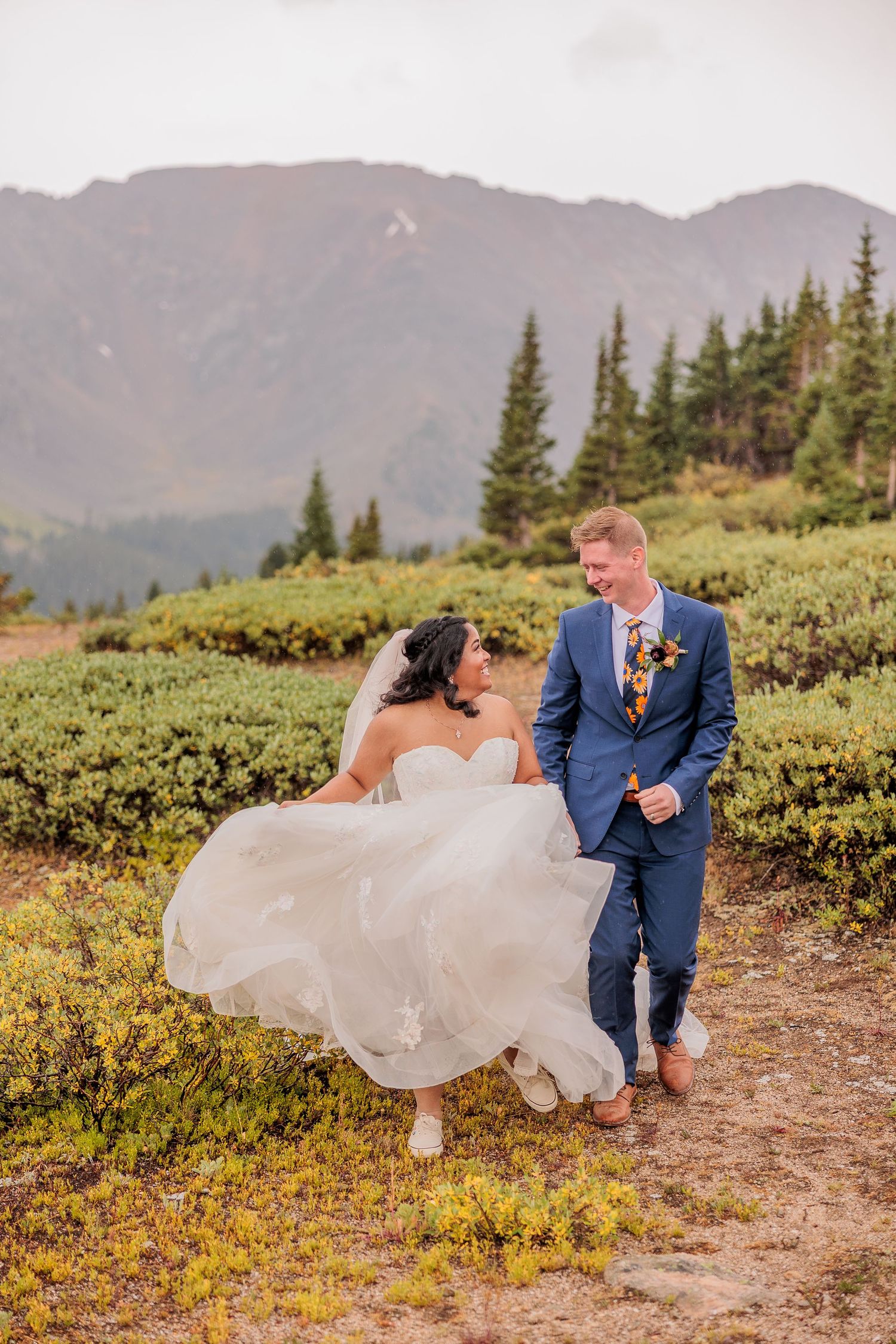 A couple in wedding attire dances playfully in a mountain meadow with evergreen trees and peaks in the background.