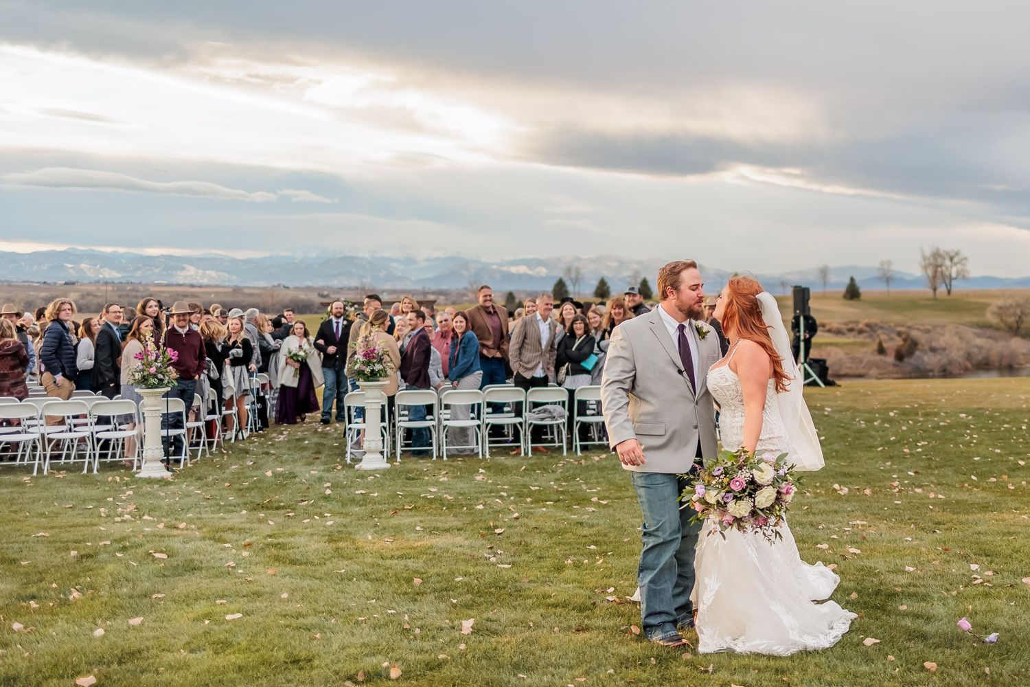 Panoramic view of an outdoor wedding ceremony with guests seated on a hilltop against mountain backdrop.