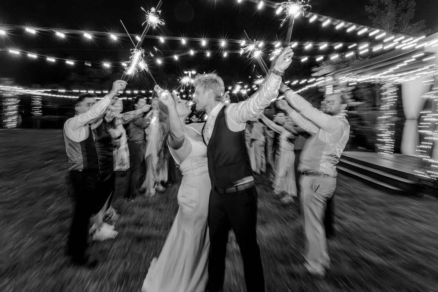 A black and white wedding reception photo with sparklers and string lights creating magical ambiance.