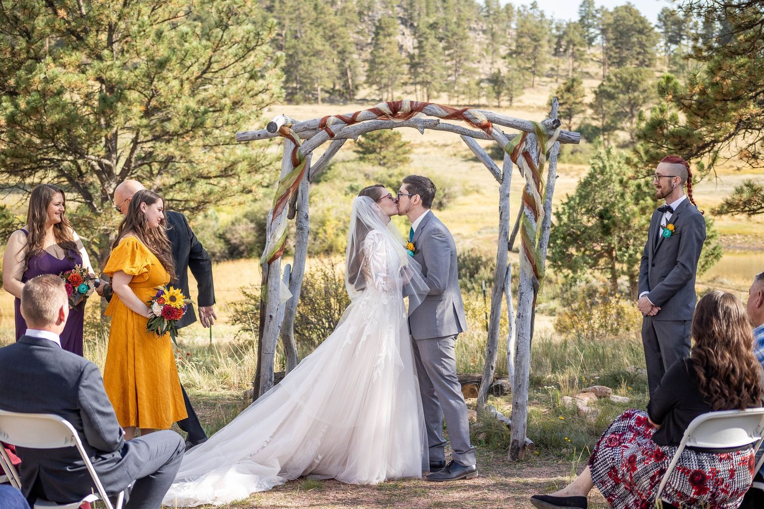 A wedding ceremony takes place in a scenic outdoor setting among tall pine trees and natural landscapes.