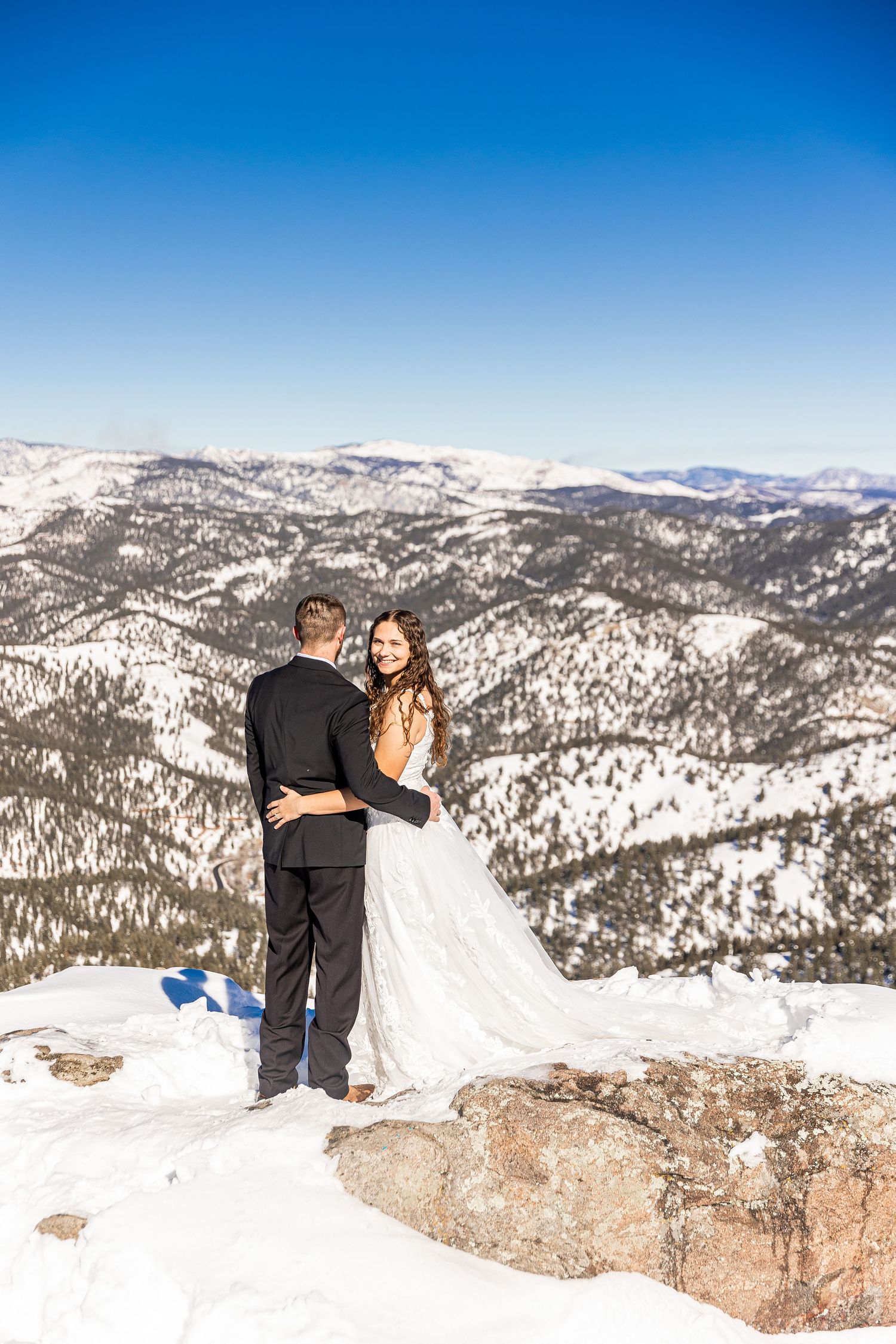 A couple in wedding attire stands at the edge of a snowy mountain overlook with snow-capped peaks in the distance.