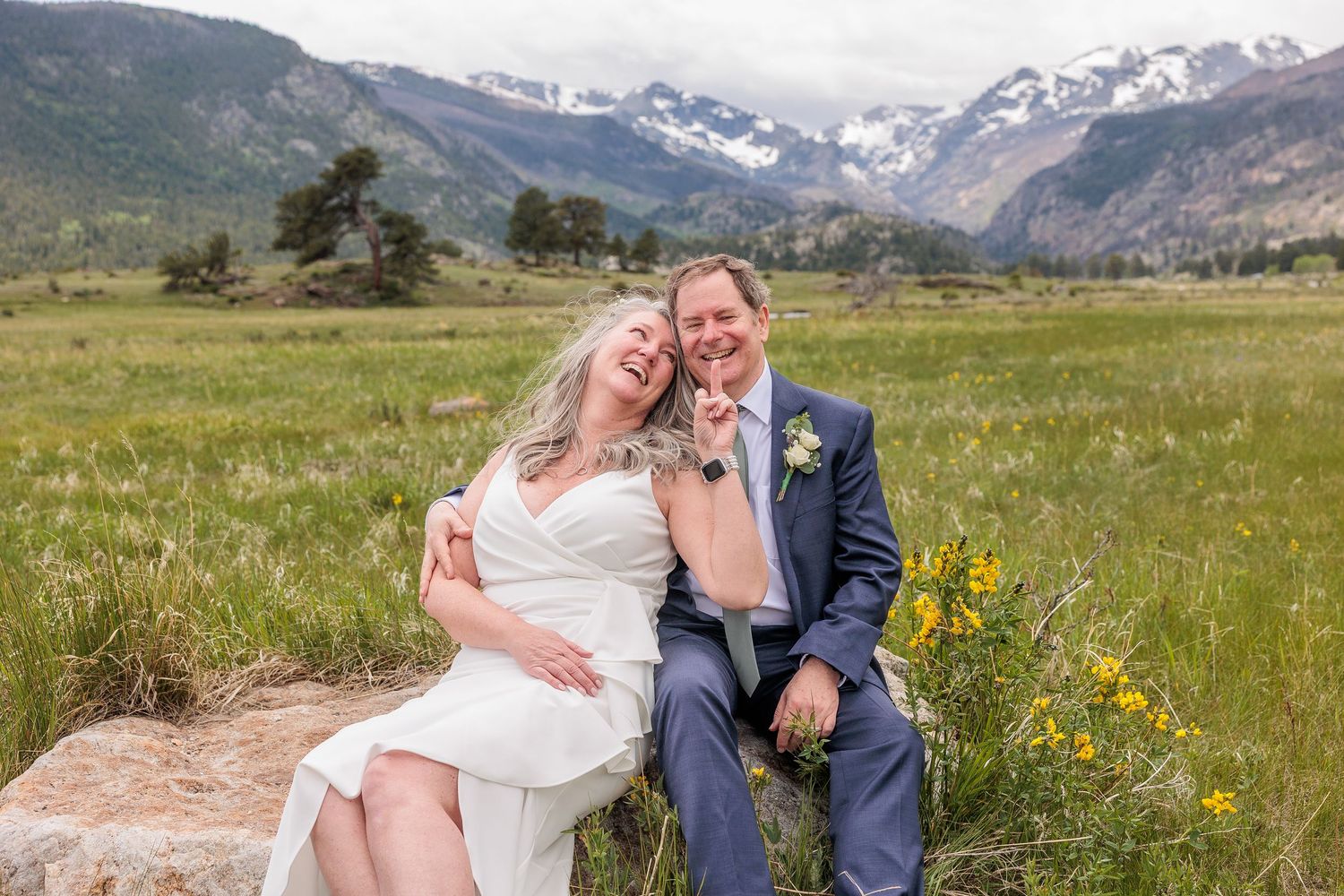 A couple embraces in a meadow with majestic Rocky Mountains and wildflowers creating a stunning backdrop.