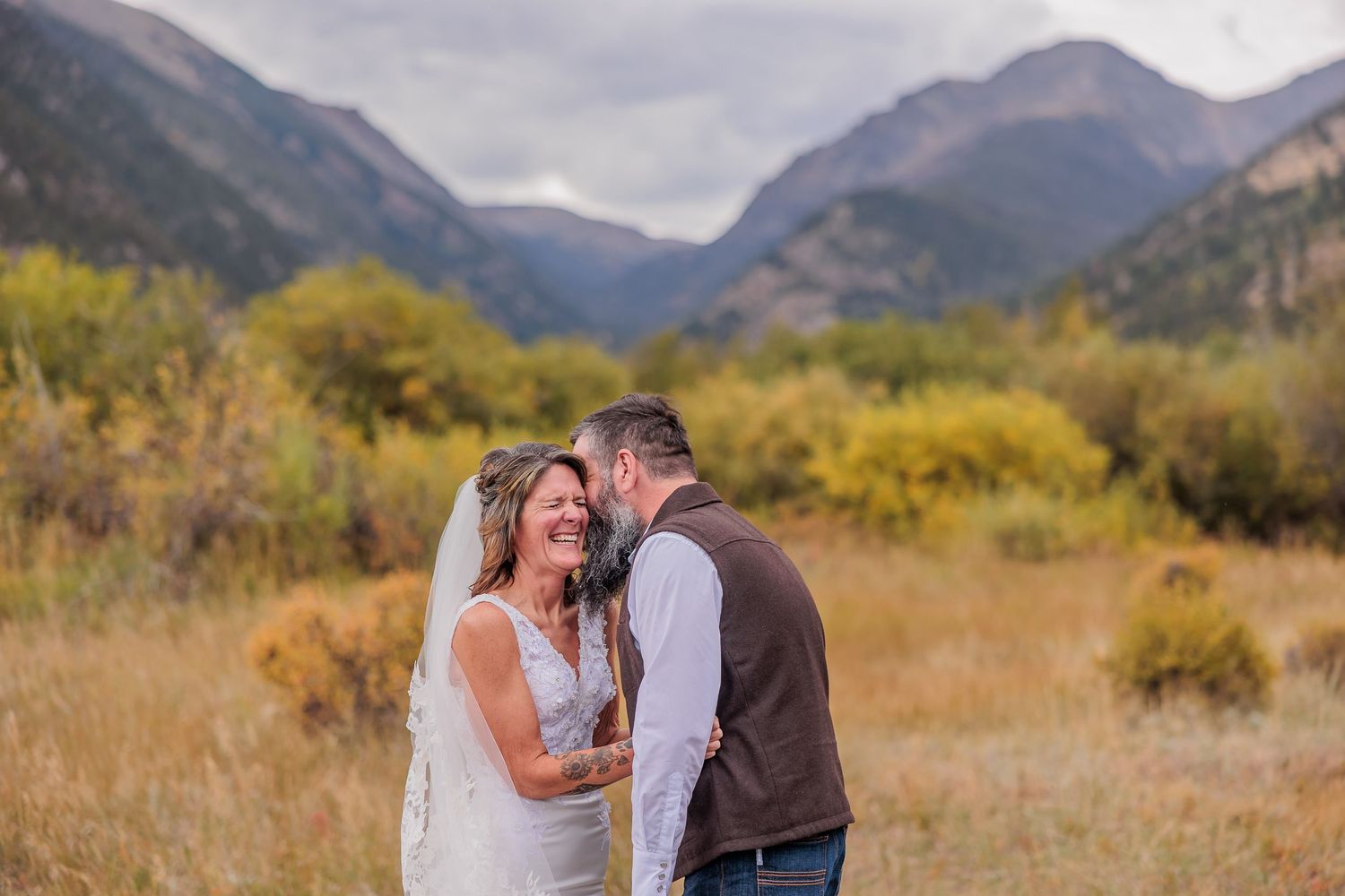 A couple shares an intimate moment in a golden meadow with majestic mountain peaks rising in the background.