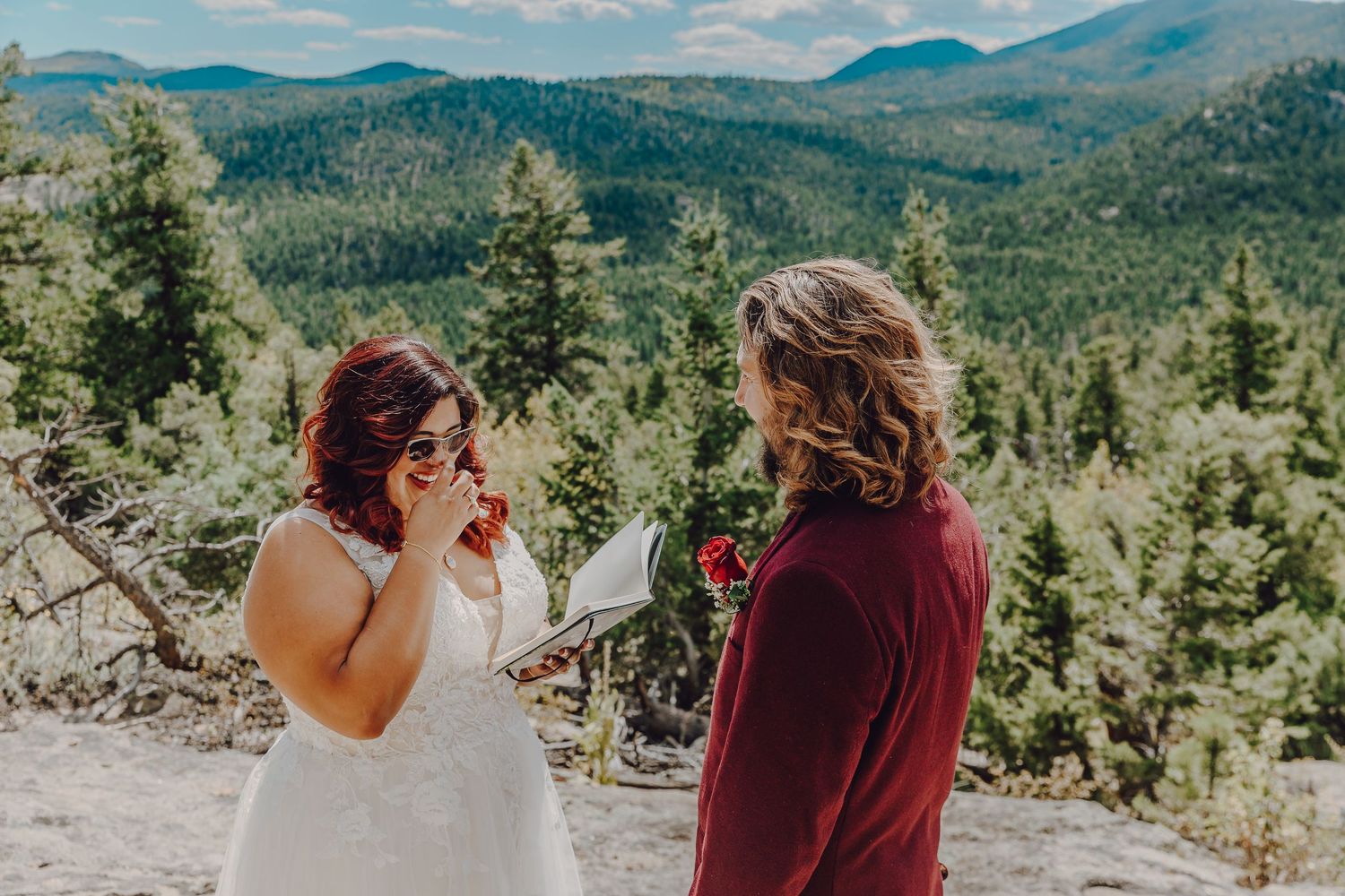 Two people share a joyful moment overlooking a scenic mountain landscape with evergreen forests and blue skies.