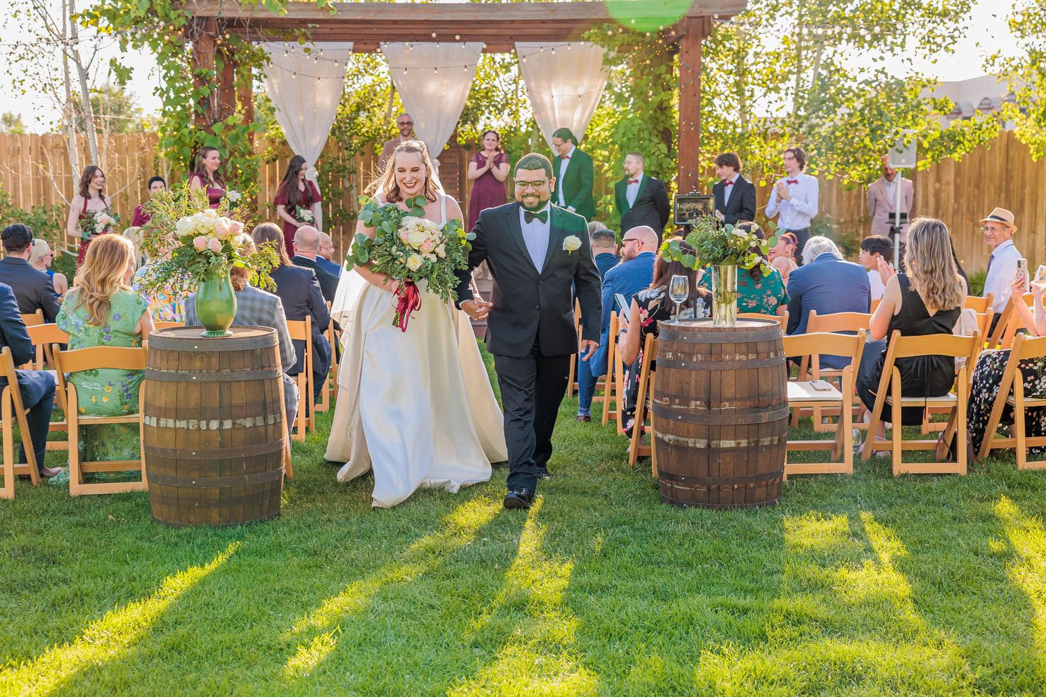 Bride and groom walk hand-in-hand down the aisle at an outdoor garden wedding ceremony with barrel decorations.