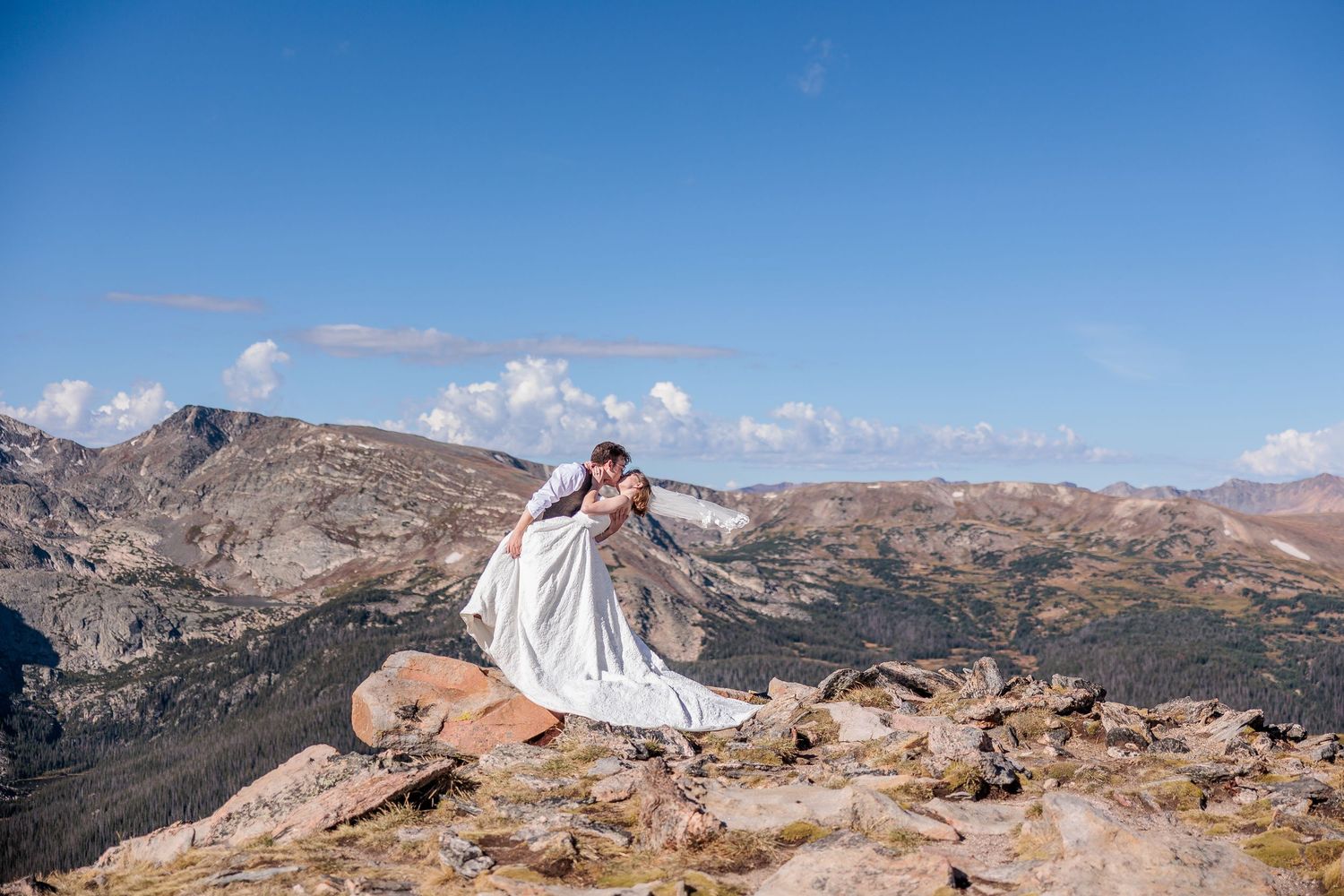 A couple in wedding attire stands on a rocky mountain peak overlooking a dramatic mountain landscape on a sunny day.