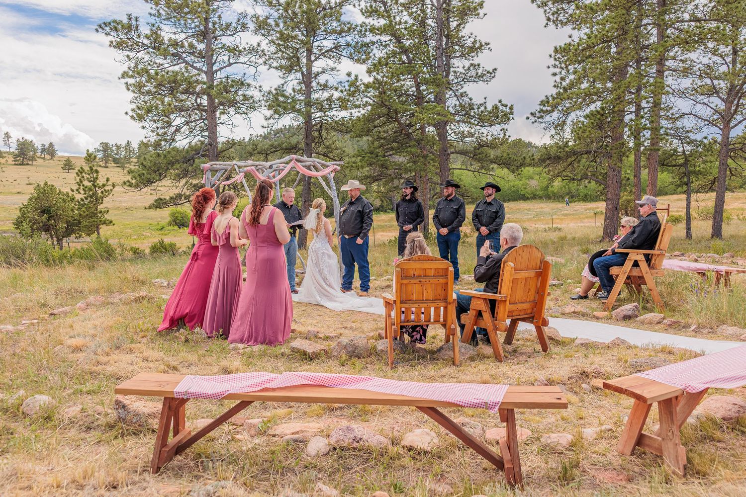 Outdoor wedding ceremony in a pine forest with bridesmaids in pink dresses and groomsmen in black attire.