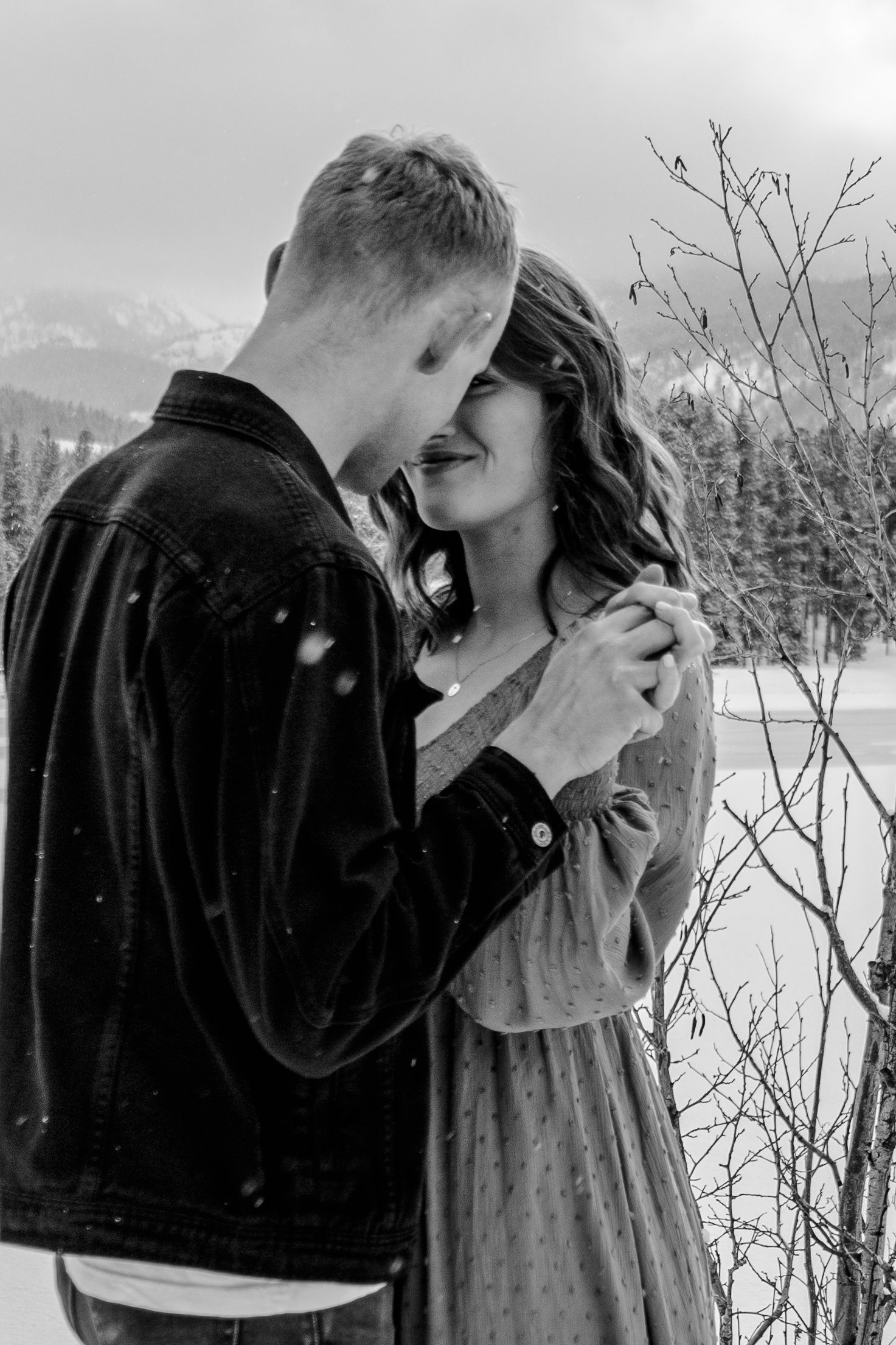 A couple shares a tender moment in a snowy winter landscape with bare trees in the background.