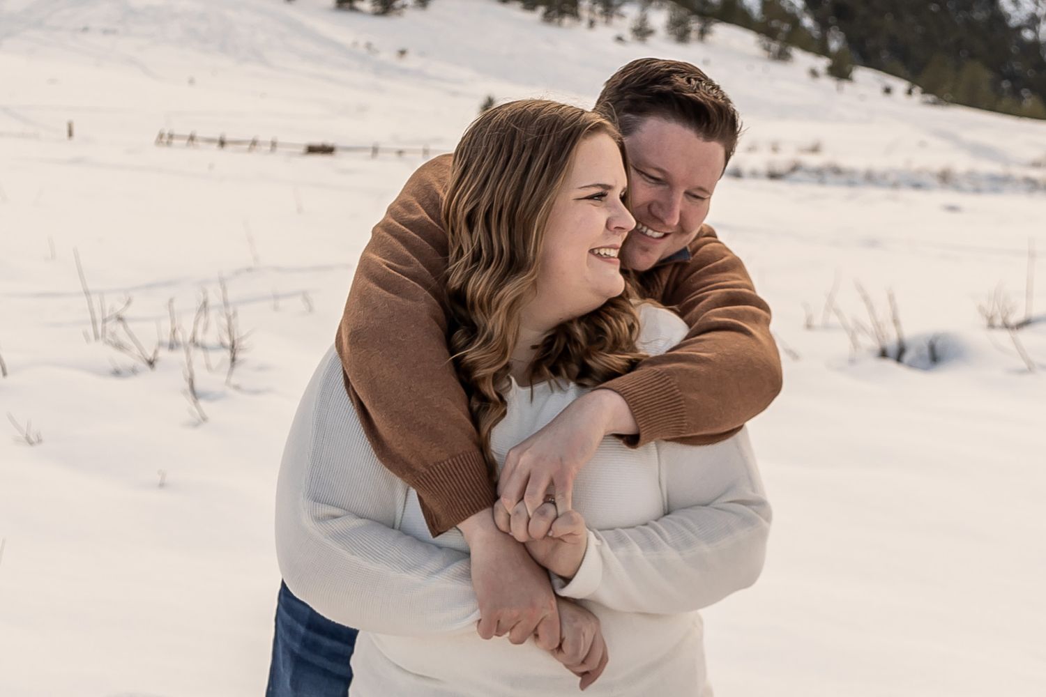 A couple in brown and white sweaters embrace lovingly in a snowy winter landscape.