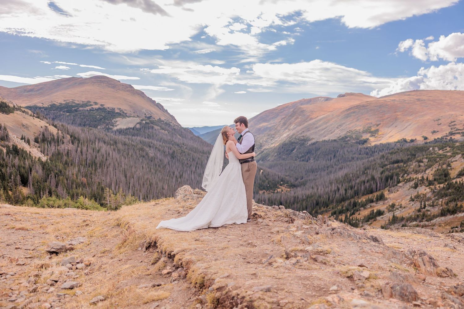 A couple in wedding attire stands atop a scenic mountain overlook with sweeping valley views in the background.