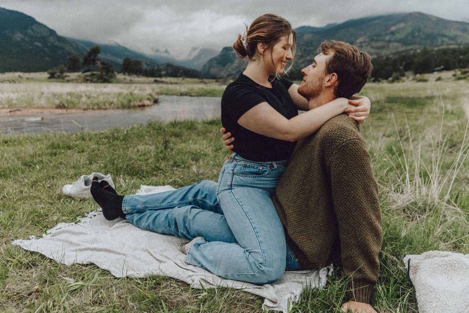 A couple shares an intimate moment while lying on a blanket in a scenic mountain meadow.