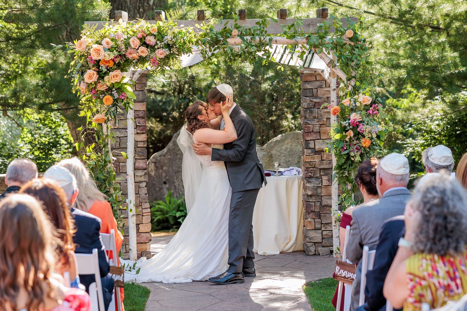 A wedding ceremony under a floral arch with guests watching the couple share their first kiss.