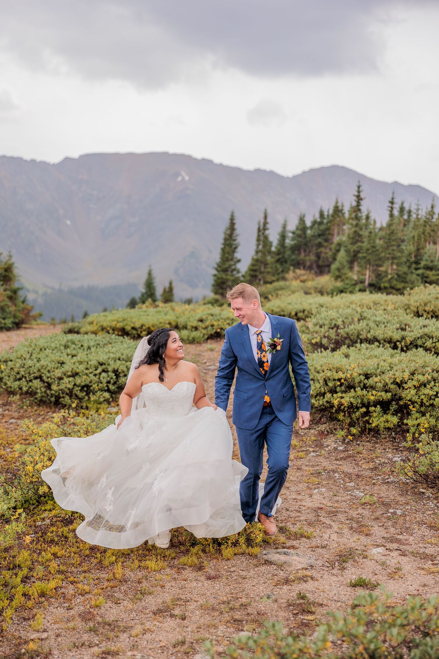 A couple in wedding attire embraces on a scenic mountain trail surrounded by evergreen shrubs and distant peaks.
