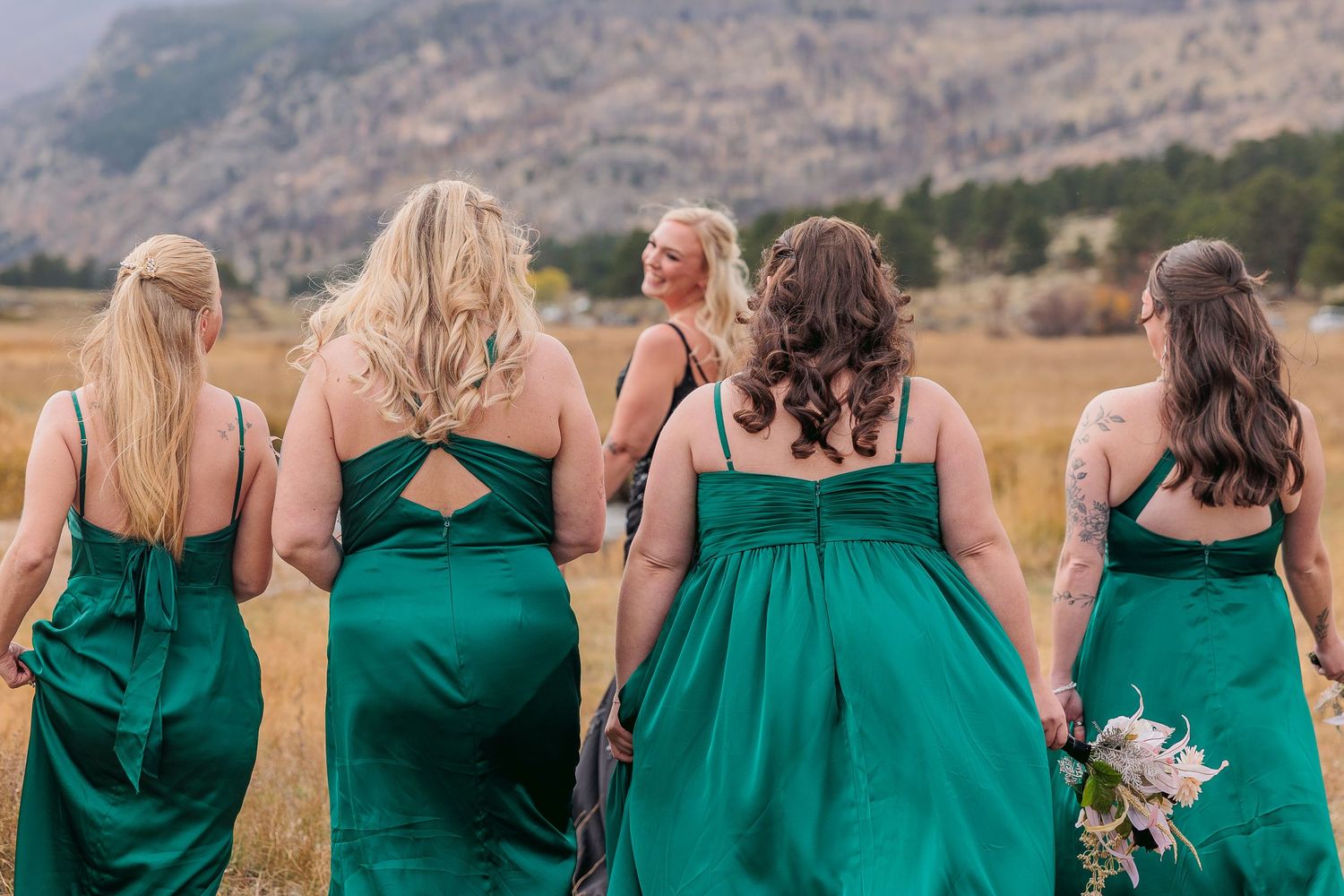 Row of bridesmaids in emerald green dresses stand with backs turned against mountain landscape backdrop.