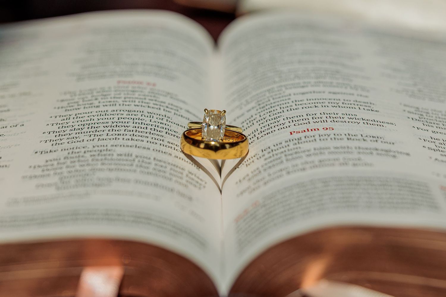 Gold wedding band and diamond engagement ring resting on open Bible pages forming a heart shape.