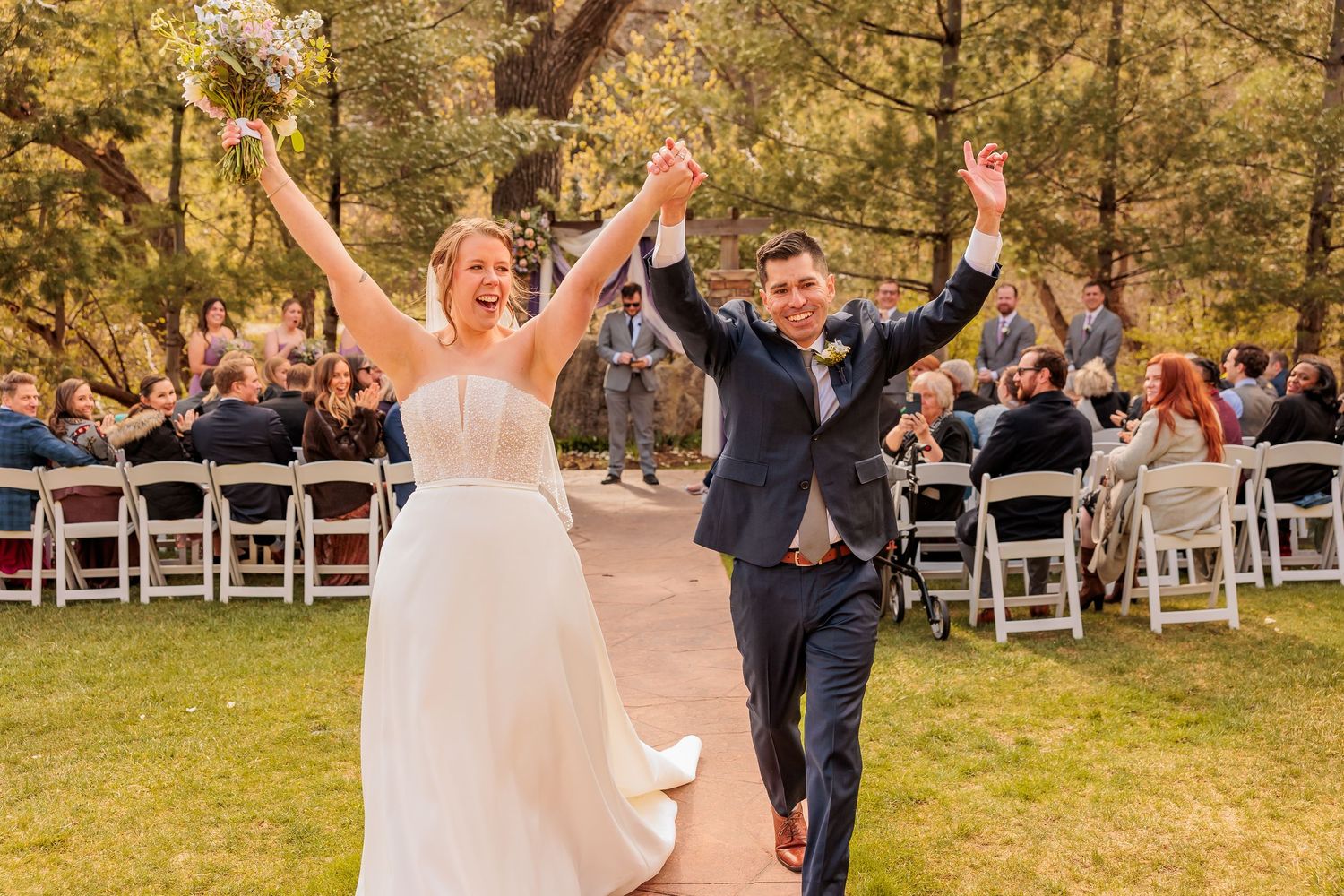 Joyful newlyweds celebrate recessional at outdoor wedding ceremony, bride in white gown and groom in navy suit raising hands.