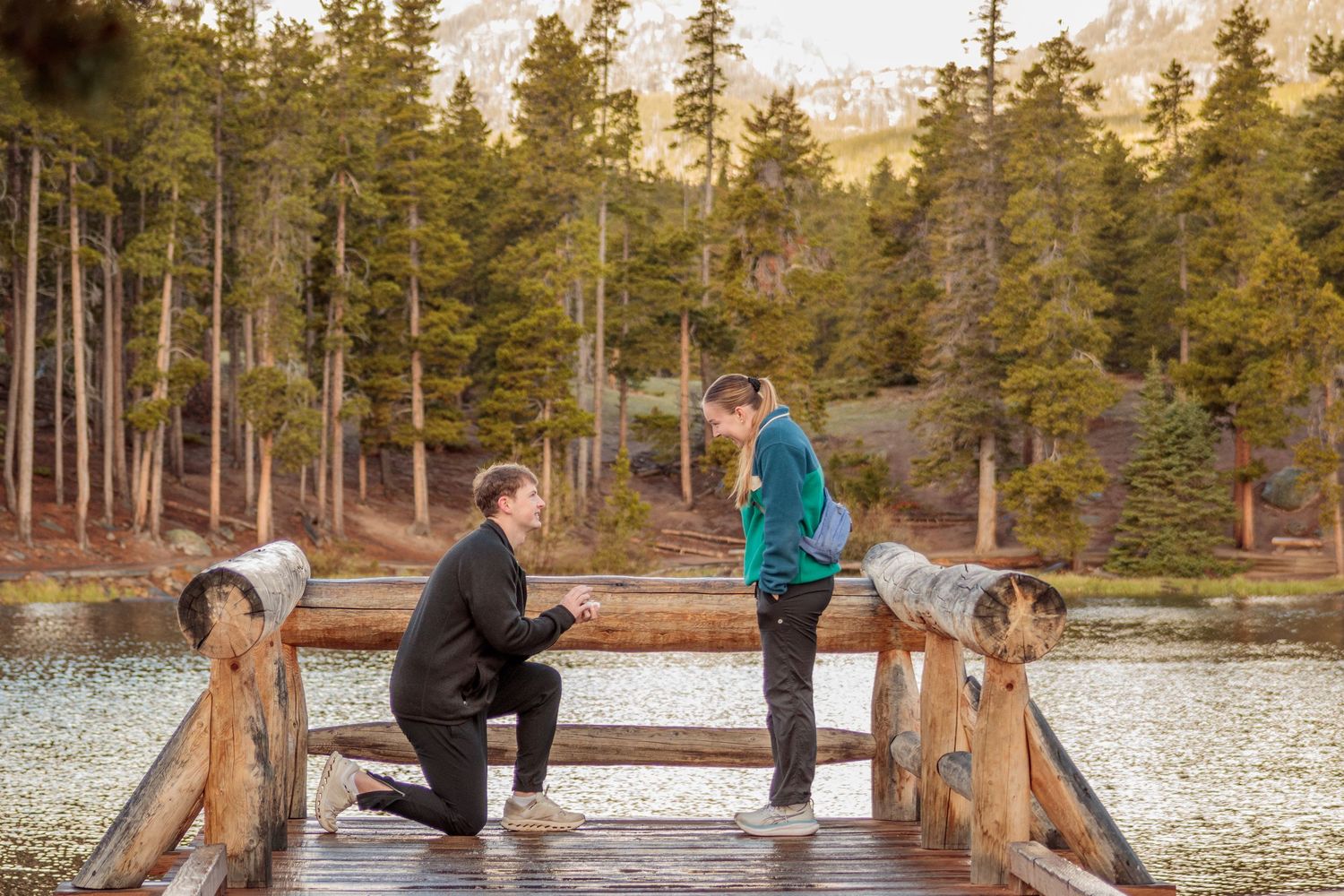 Man proposing to woman on wooden dock by mountain lake surrounded by pine trees and scenic peaks.
