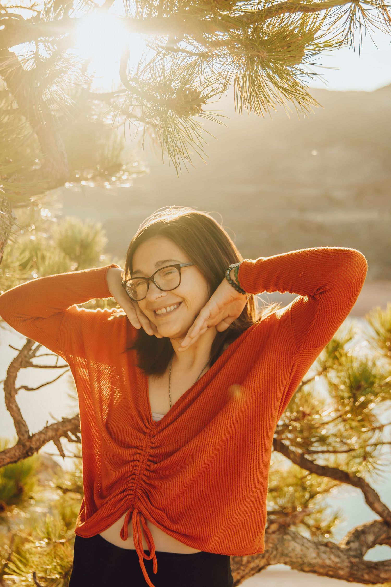 Person in orange sweater and glasses enjoying golden sunlight through pine trees on a warm afternoon.