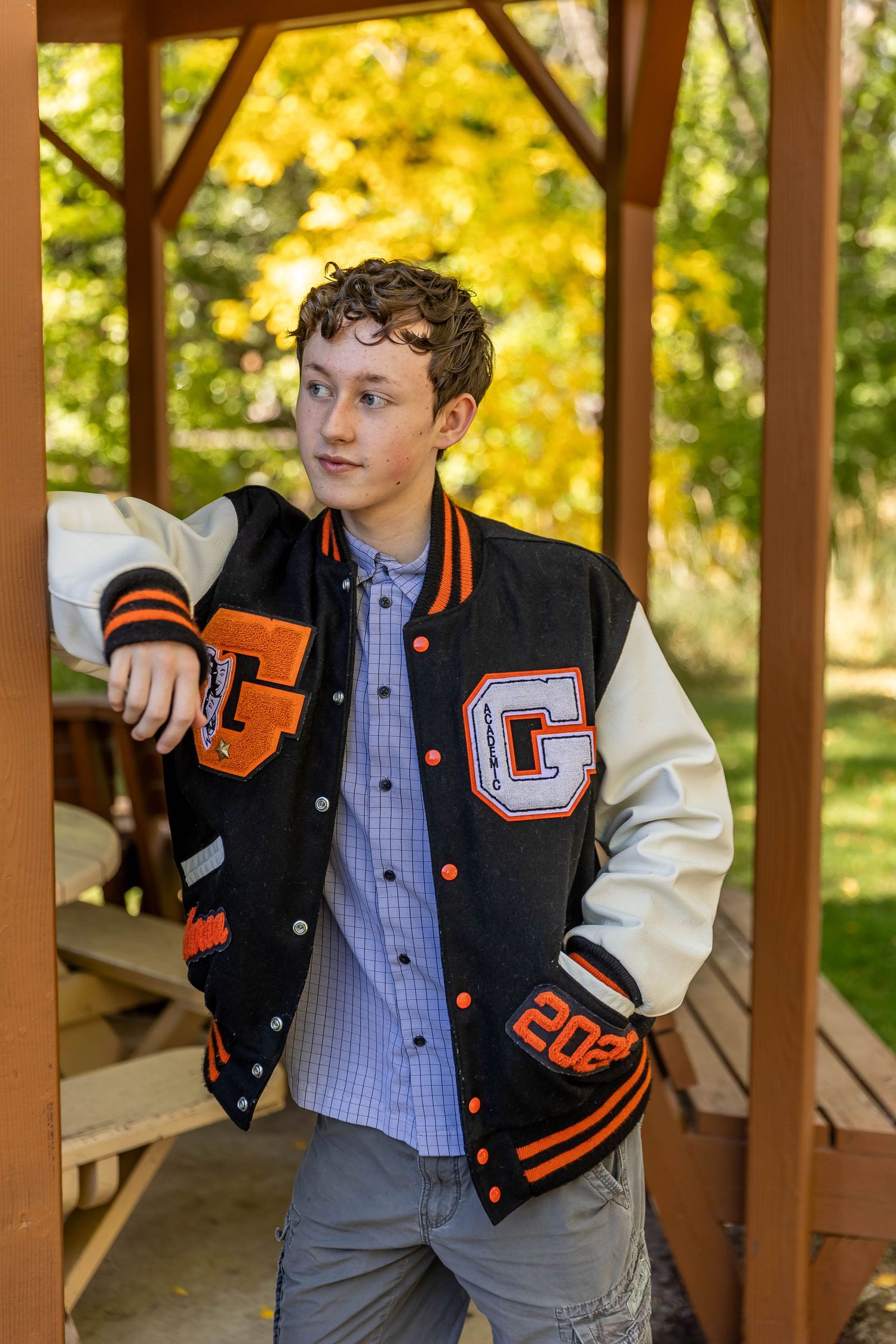 Student wearing a black and orange varsity letterman jacket poses casually in a wooden gazebo on a sunny autumn day.