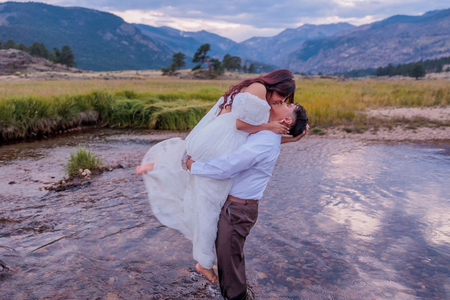 A couple shares a romantic moment in a flowing river surrounded by mountain scenery at sunset.