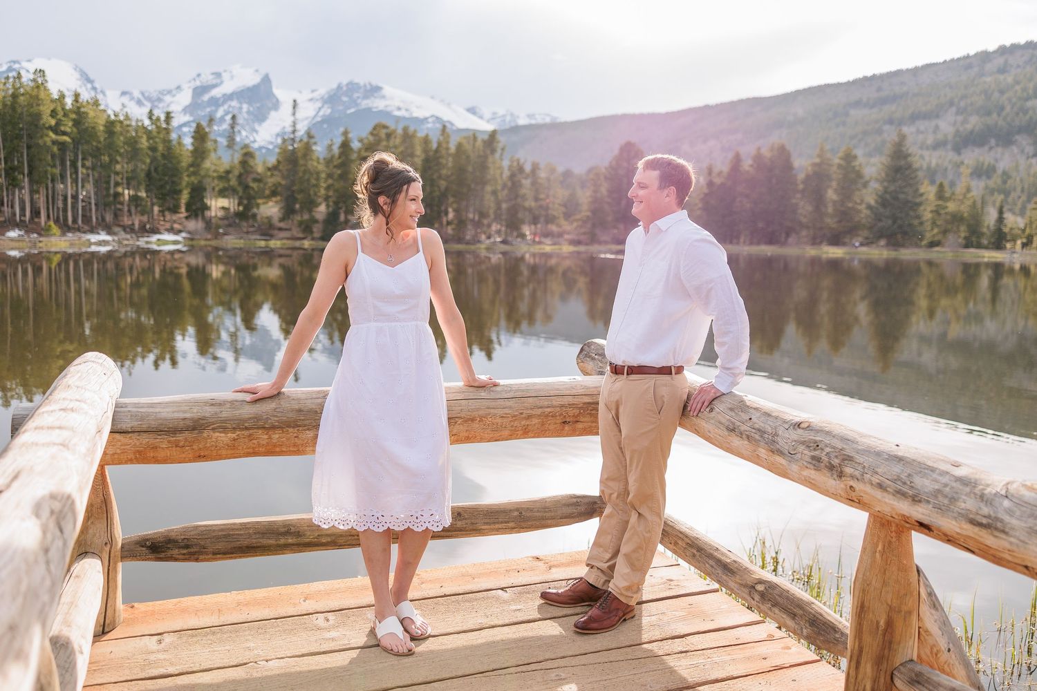 Couple sits together on rustic wooden dock overlooking serene mountain lake during sunset.