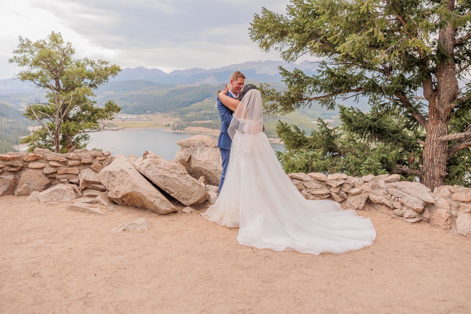 A couple in wedding attire stands on a rocky mountain overlook with scenic mountain vistas in the background.