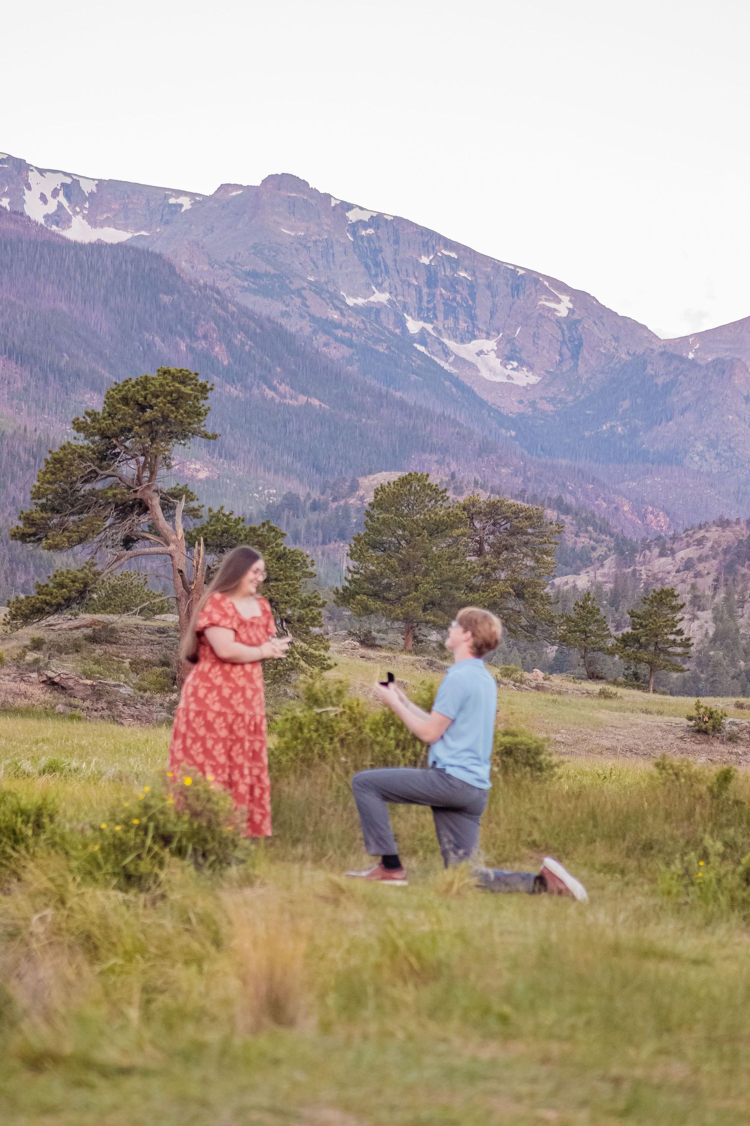 Man proposing on one knee to woman in red dress with stunning Rocky Mountain backdrop at sunset.