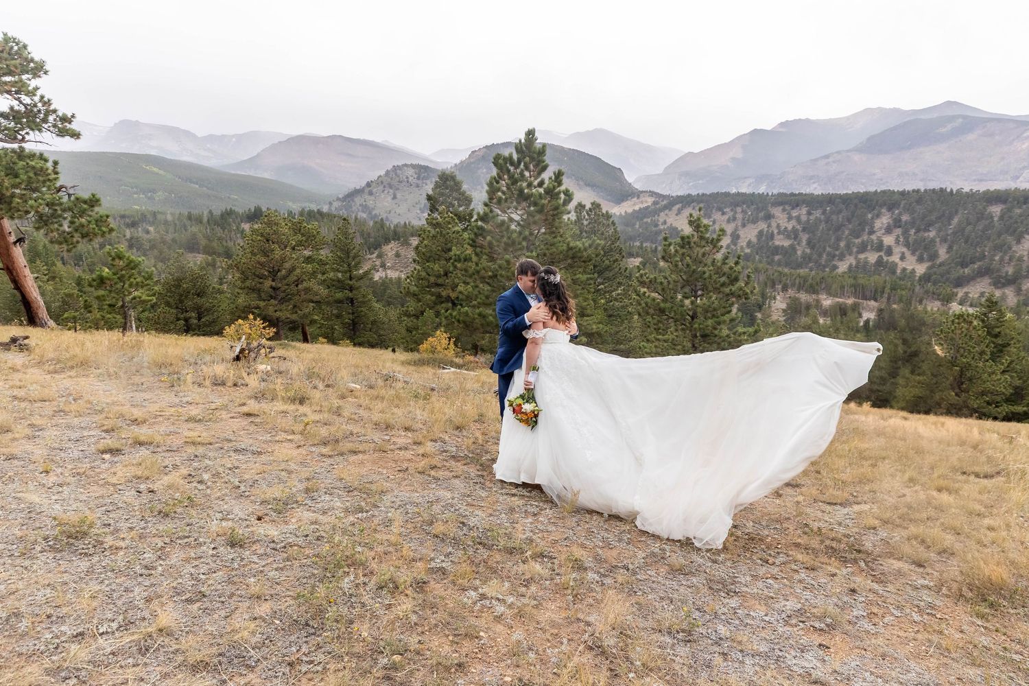 A wedding dress train flows in the wind against a scenic mountain landscape backdrop during a photo session.